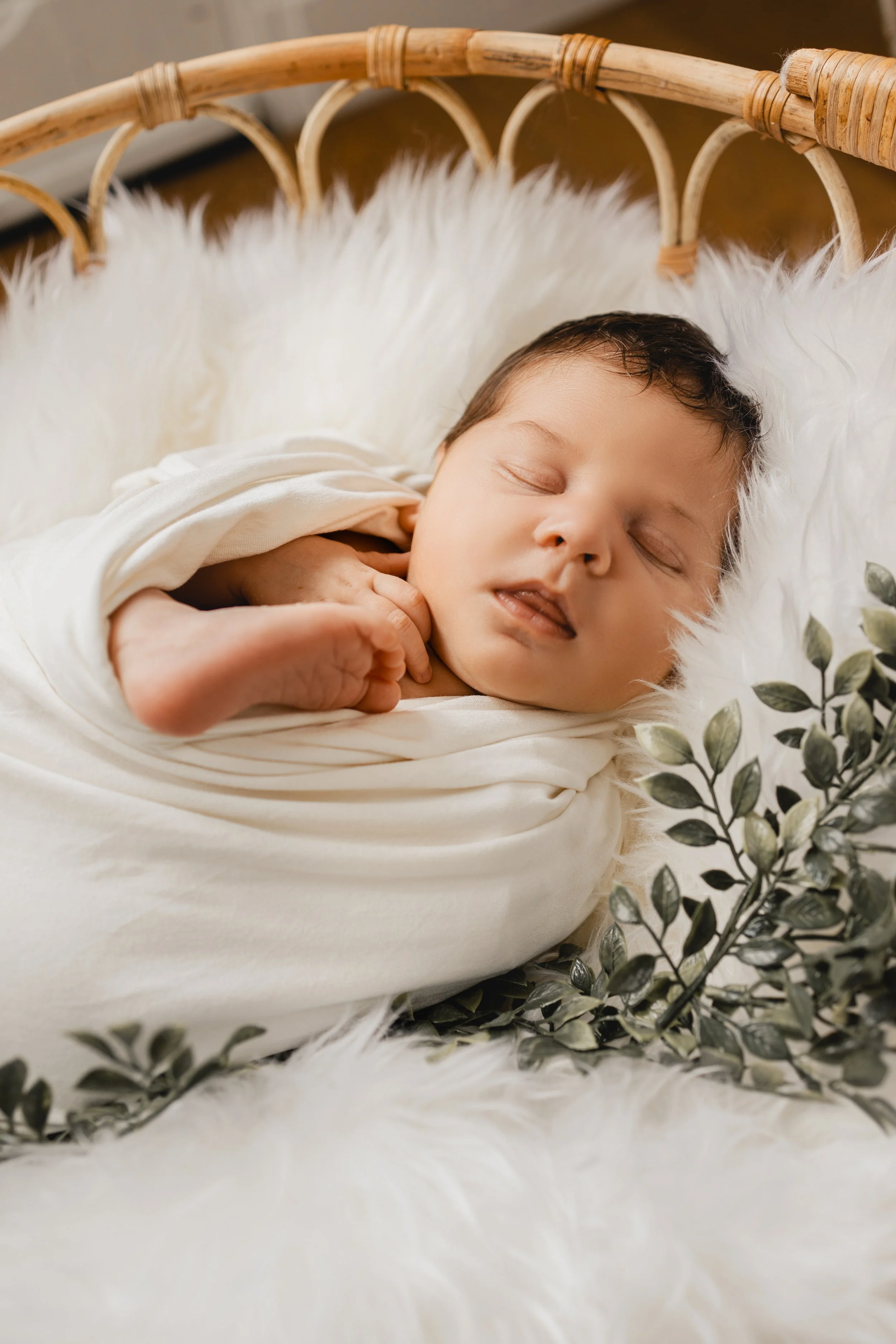 Un bébé dormir enroulé dans un drap blanc sur un lit avec une couverture en fourrure et des feuilles vertes autour.