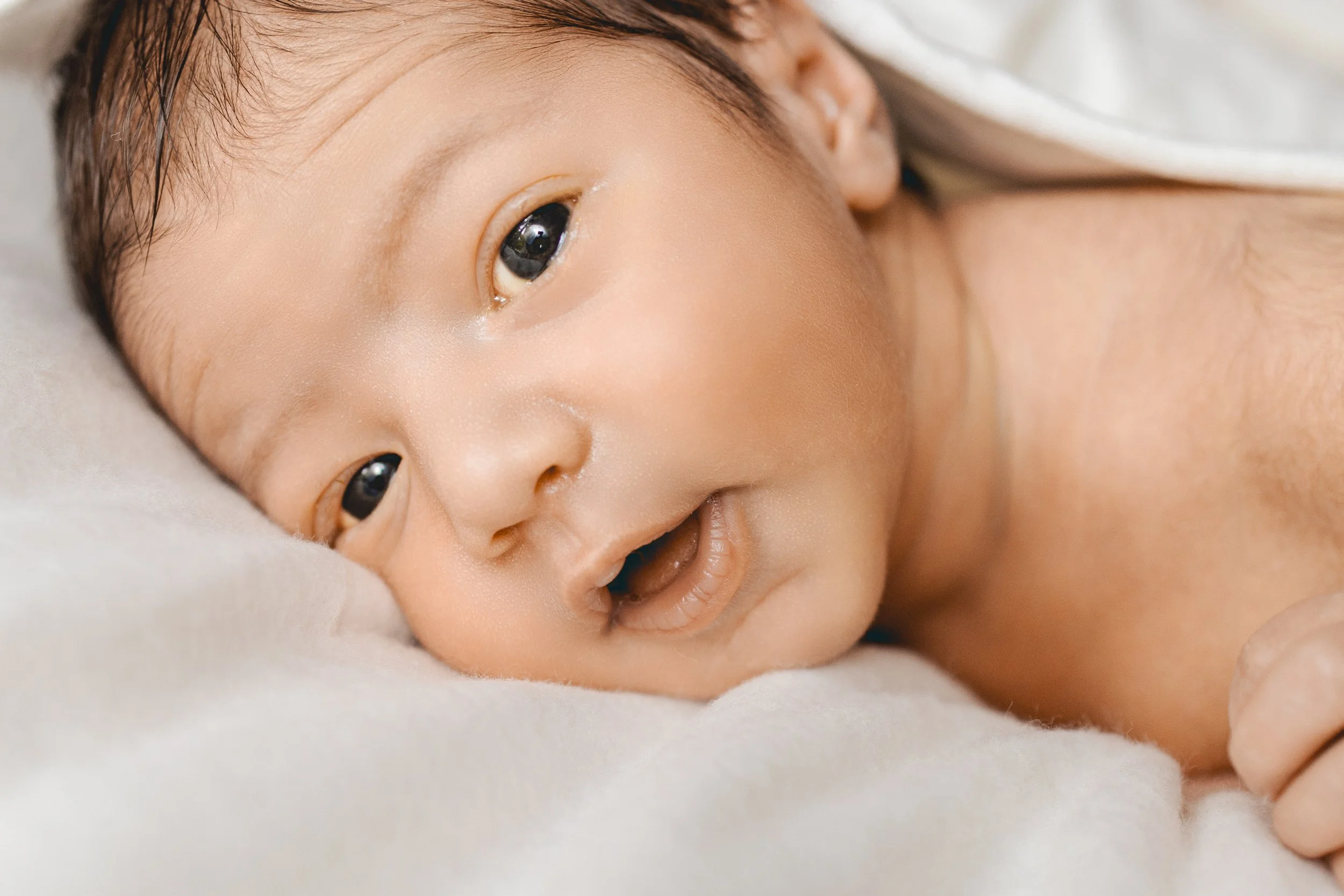 Un bébé avec la peau claire, allongé sur un lit, regardant vers l'objectif, avec une expression ouverte et curieuse.