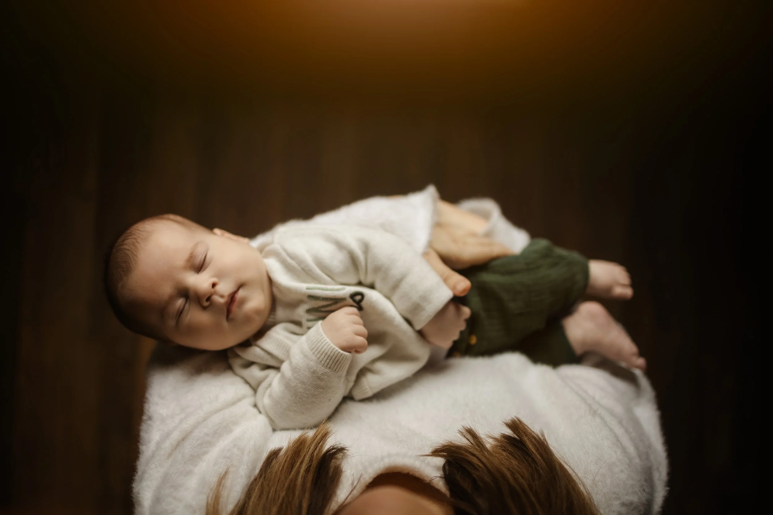 Un bébé dormant allongé sur une personne, avec des mains posées doucement sur lui, dans un environnement chaud et sombre.