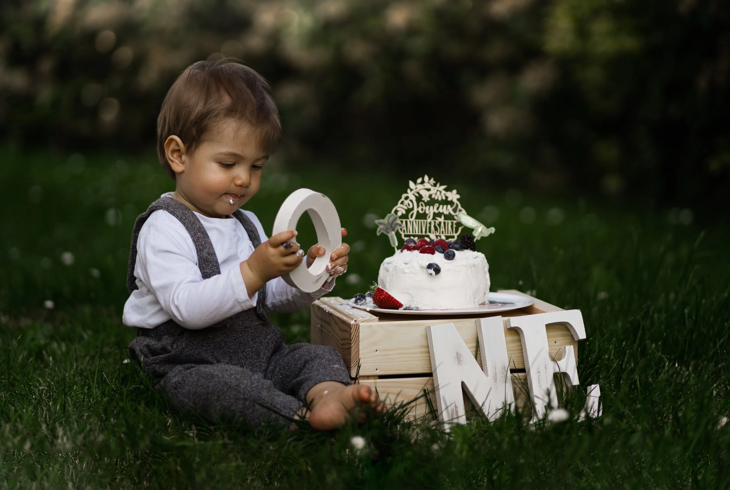 Un petit garçon assis dans l'herbe devant un gâteau d'anniversaire avec des fruits, avec un décor de fête, dans un jardin. Il tient un chiffre '0' en bois blanc et regarde le gâteau.