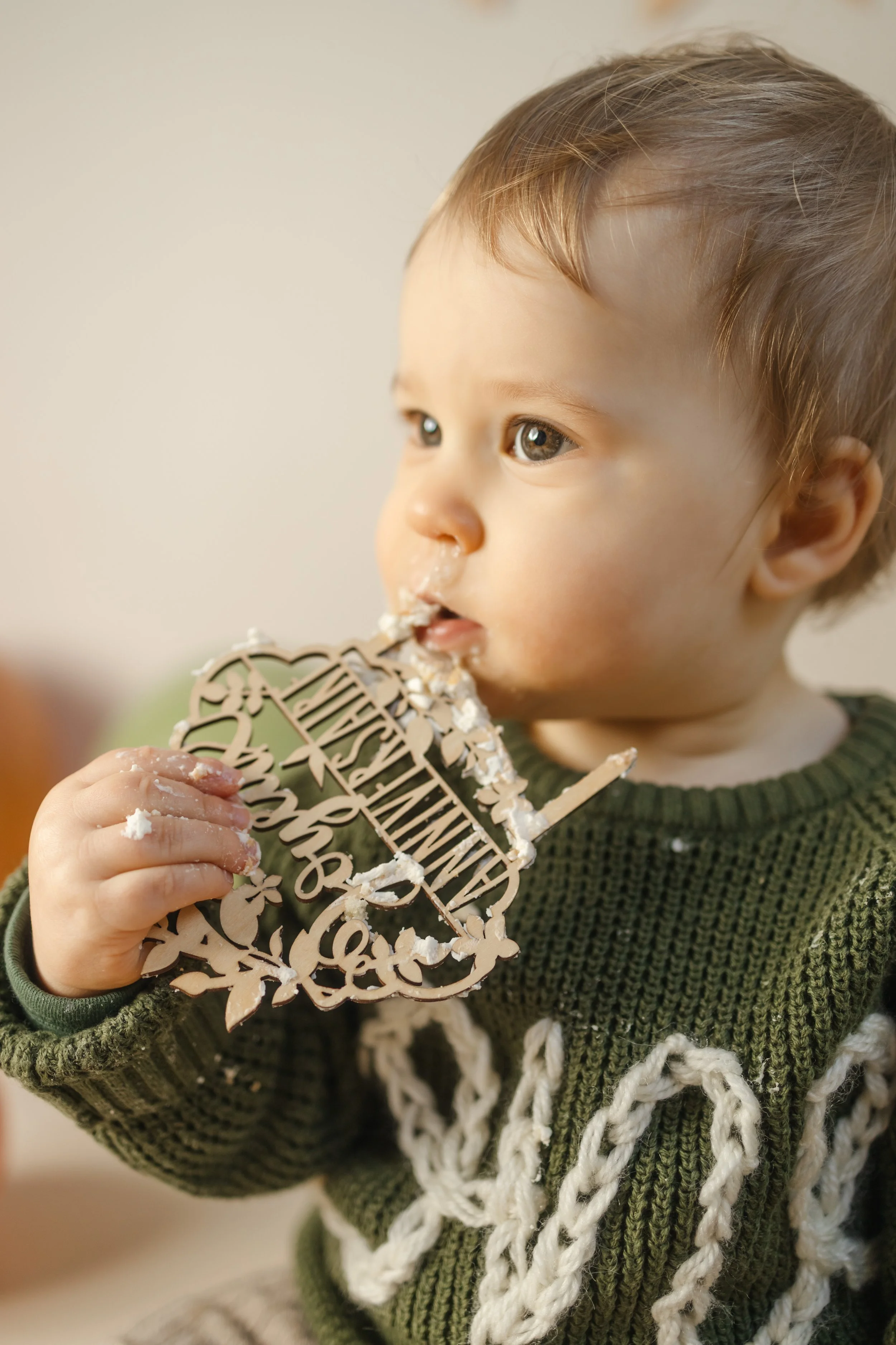 Un jeune enfant mangeant de la crème ou du gâteau, tenant un ornement en bois avec des mots en français, en portant un pull vert avec un motif torsadé blanc.