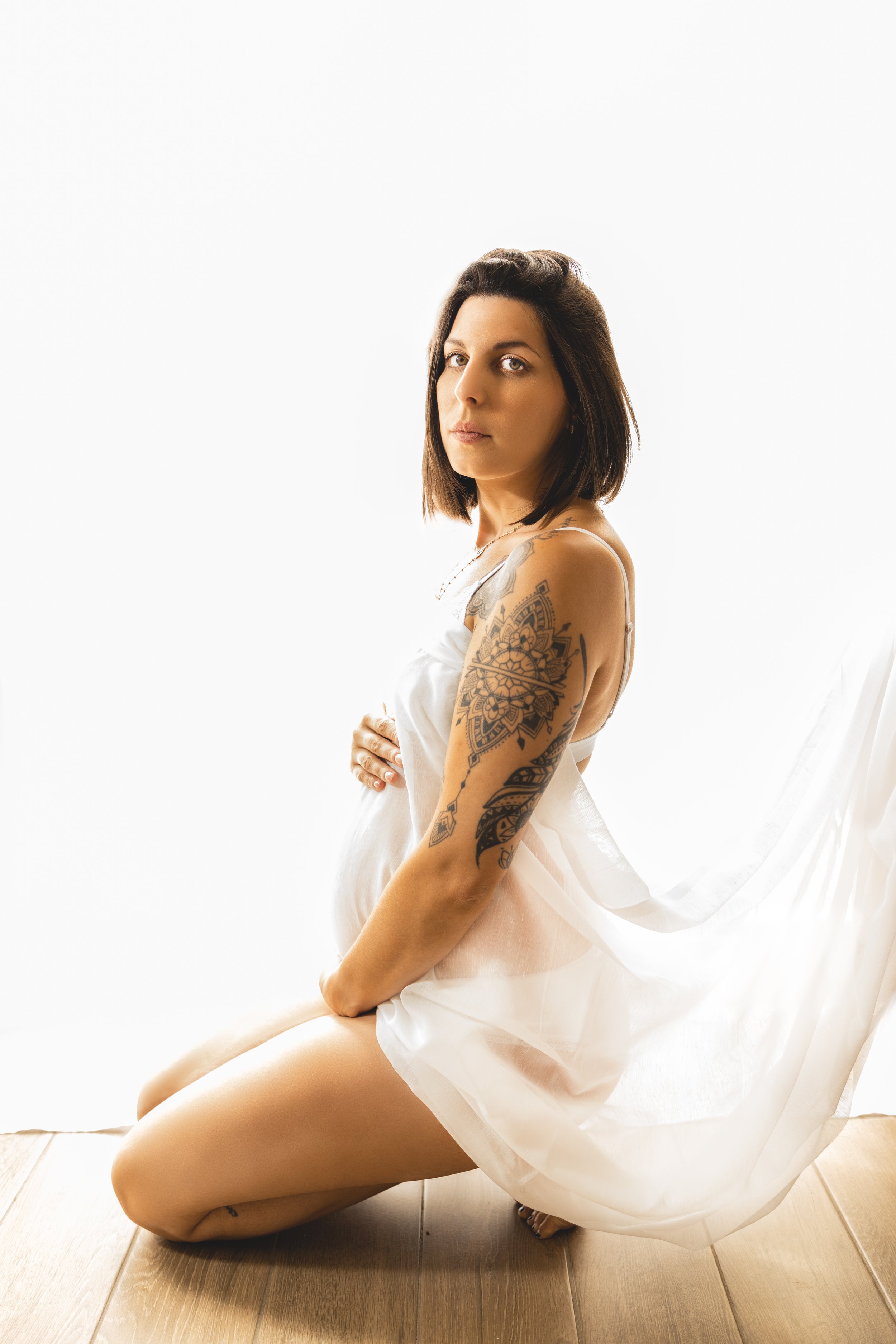 Femme enceinte en robe blanche, assise à genoux sur un parquet, avec tatouages sur le bras gauche et l'épaule droite, regardant vers l'objectif, fond blanc lumineux