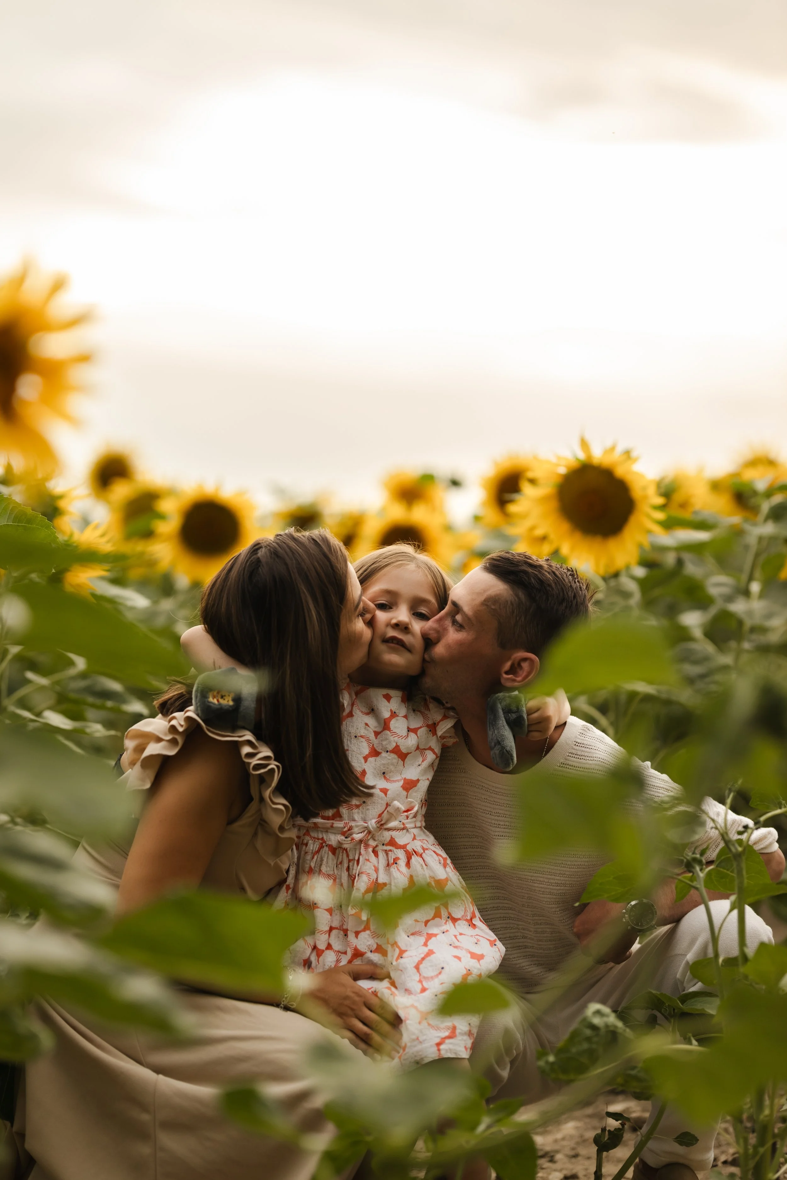 Une famille composée d'une mère, d'un père et d'une jeune fille, partageant un moment affectueux dans un champ de tournesols sous un ciel légèrement nuageux.