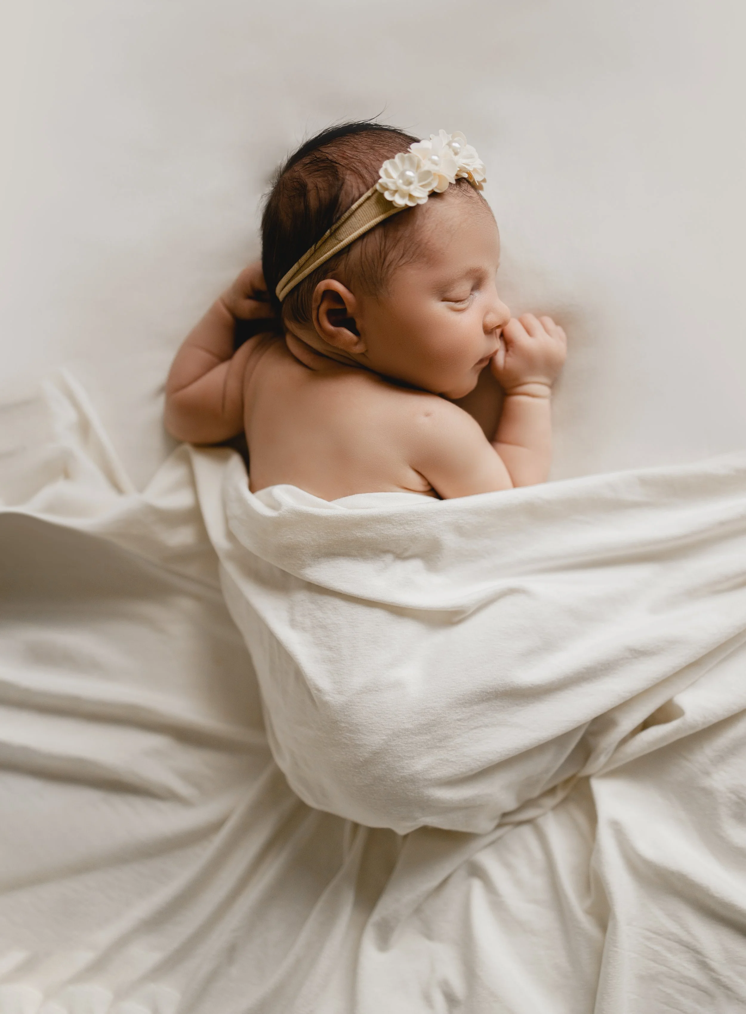 Un bébé allongé sur un lit, endormi, avec un bandeau à fleurs sur la tête et enveloppé dans une draperie blanche.