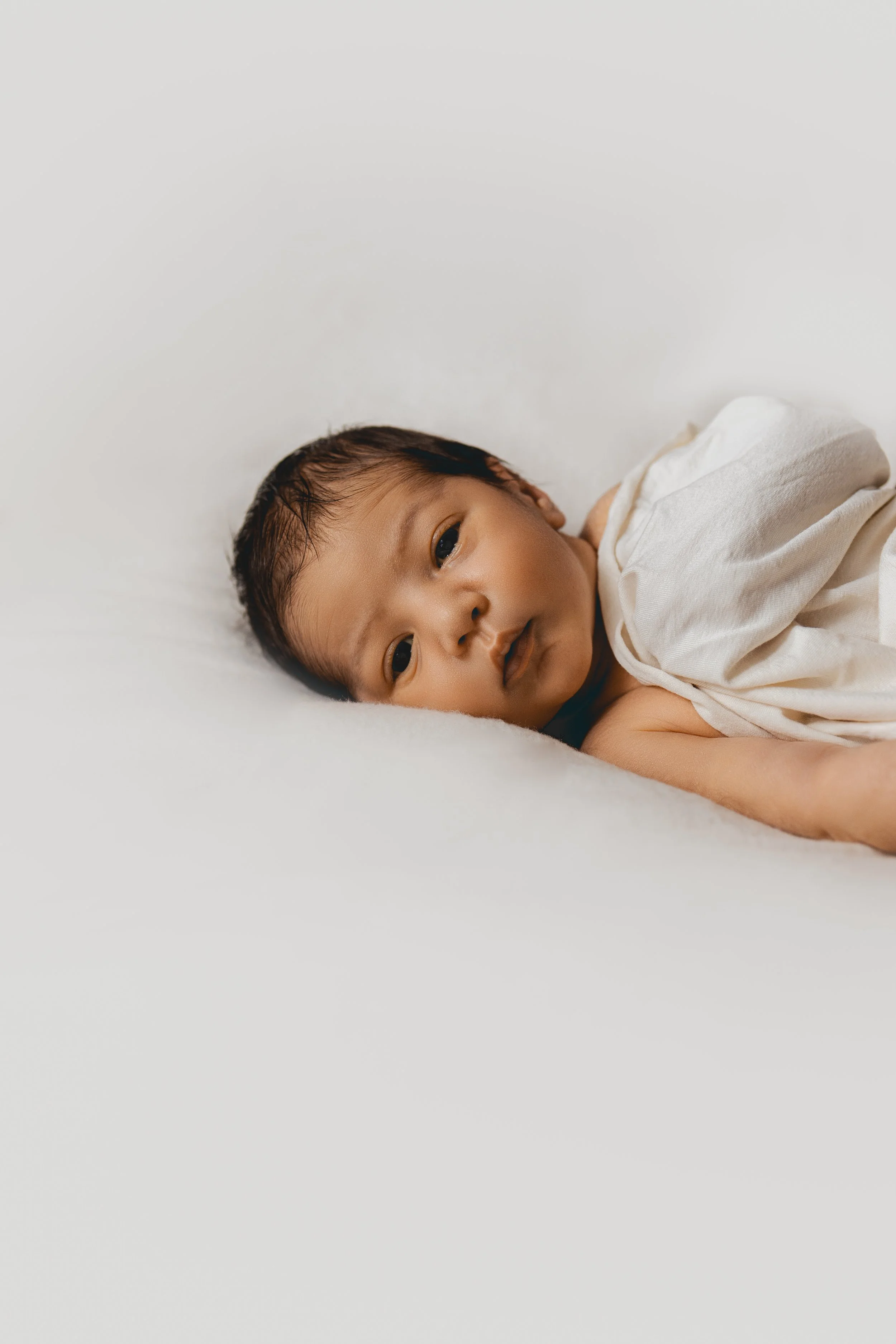 Bébé allongé sur un lit blanc, regardant vers l'objectif, avec une expression neutre. Lunettes de bébé avec une couverture blanche.