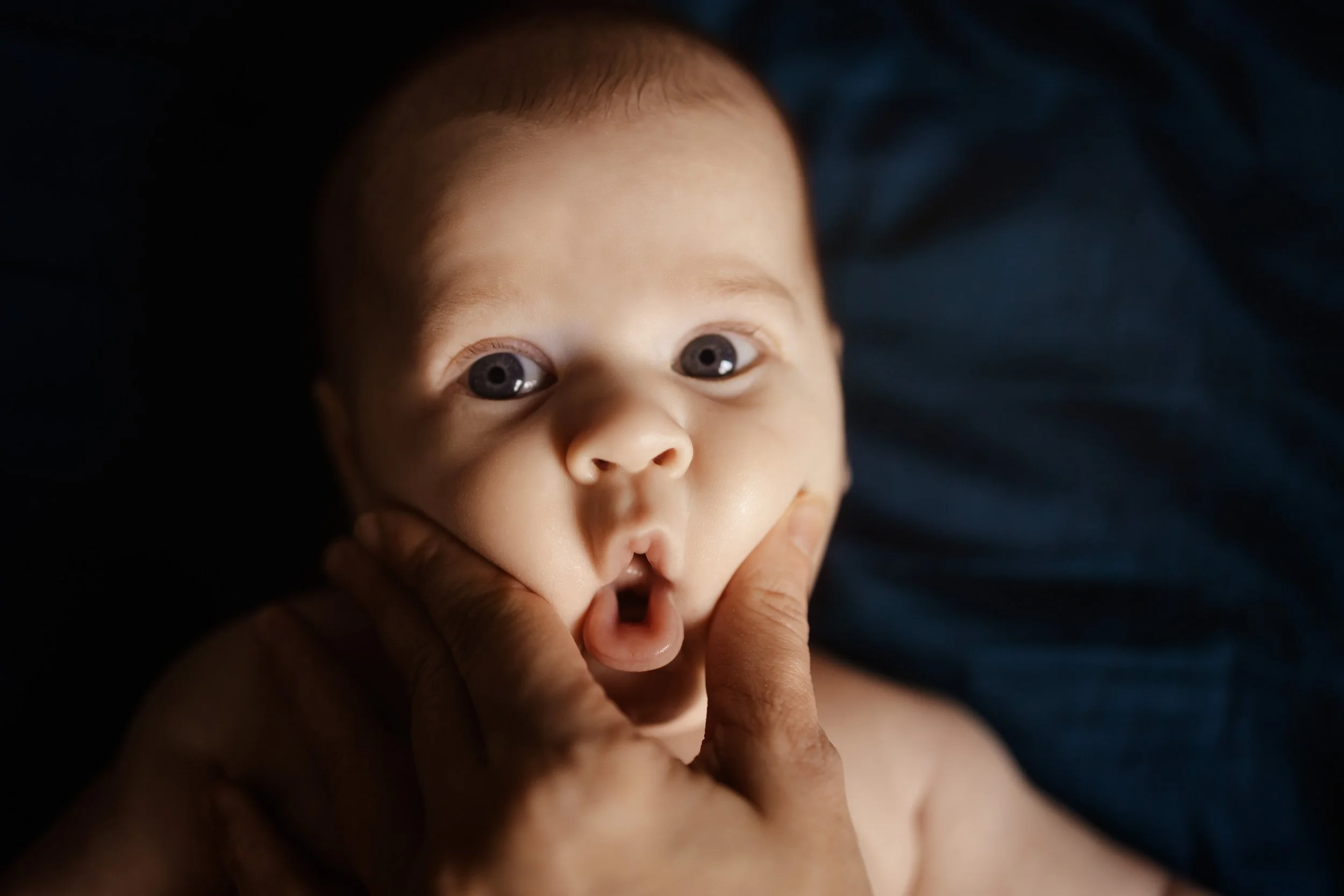 Gros plan d'un bébé avec des yeux grands ouverts, une main tenant son visage, oreilles en relief, fond sombre.