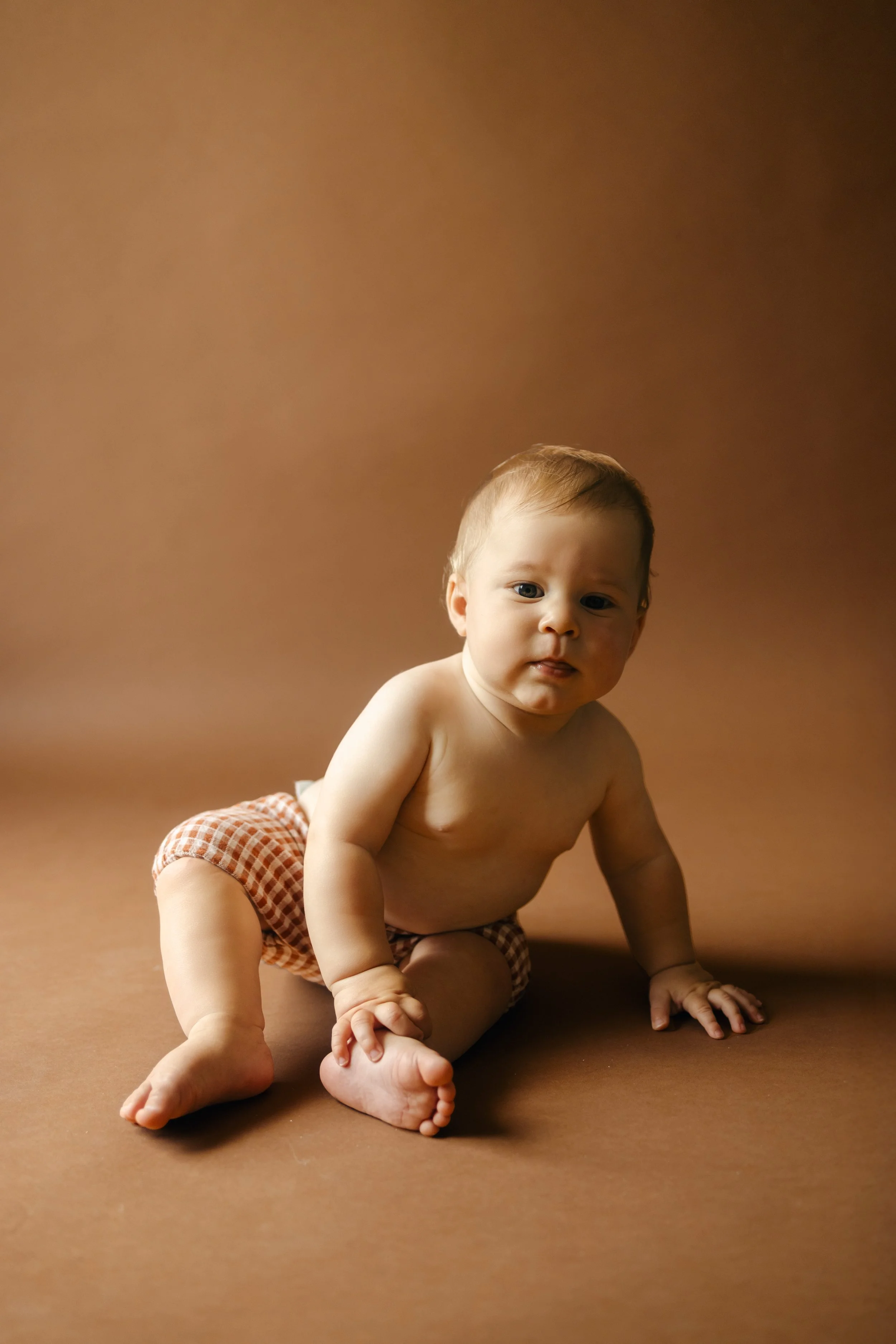 Jeune bébé à la peau claire et cheveux bruns, vêtu de couches en tissu à carreaux, assis sur un fond marron.