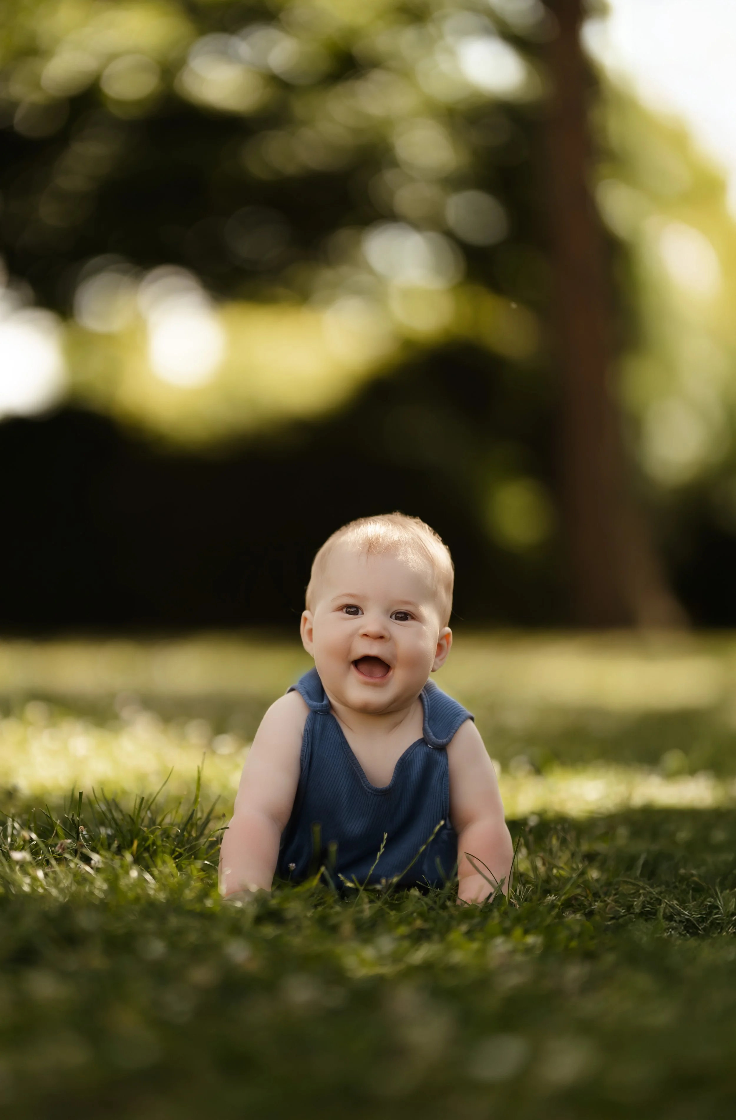 Un bébé souriant, allongé sur l'herbe dans un parc, avec des arbres en fond, en plein air par une journée ensoleillée.