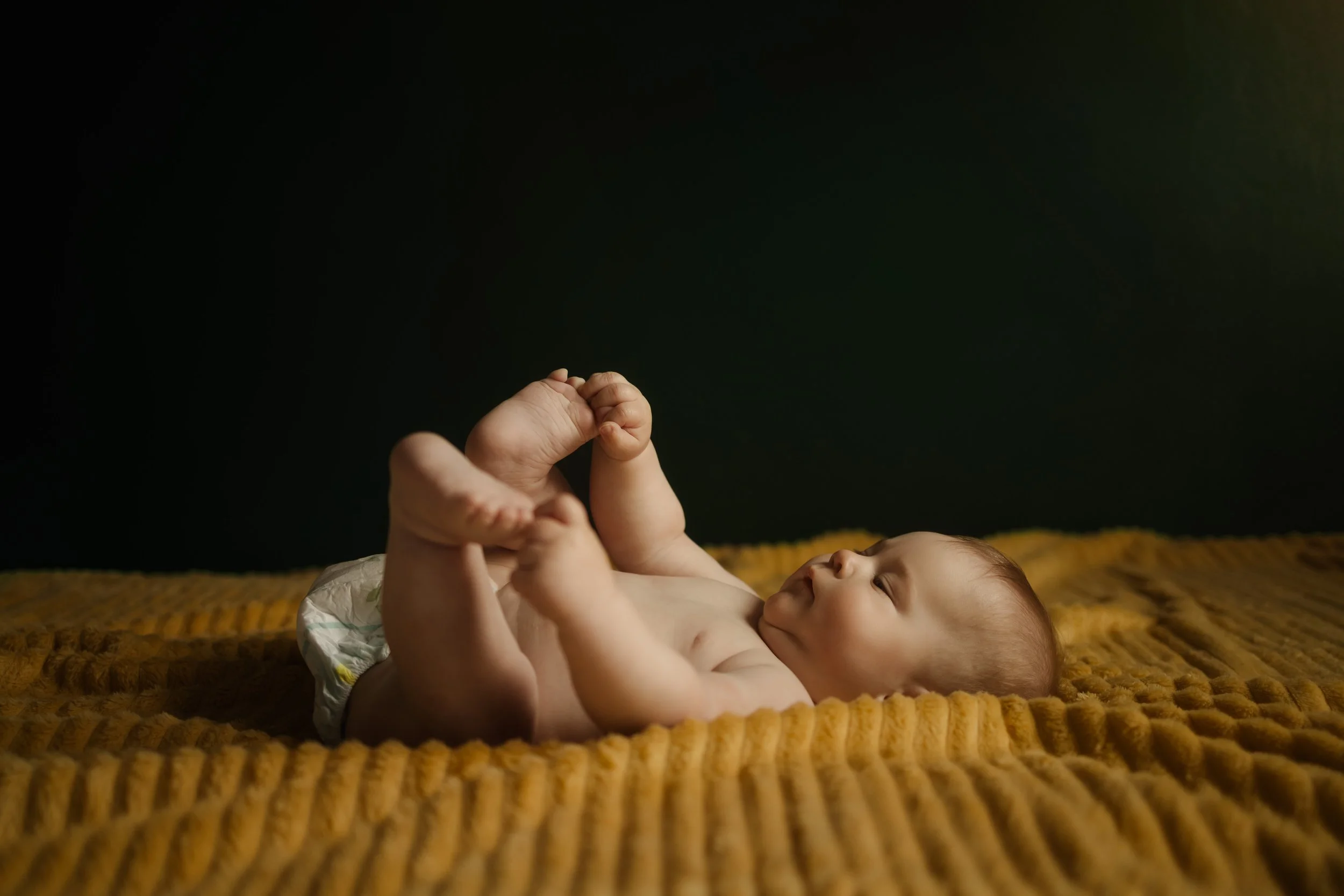 Un bébé allongé sur un lit avec une couverture jaune, souriant et jouant avec ses pieds dans un environnement sombre.