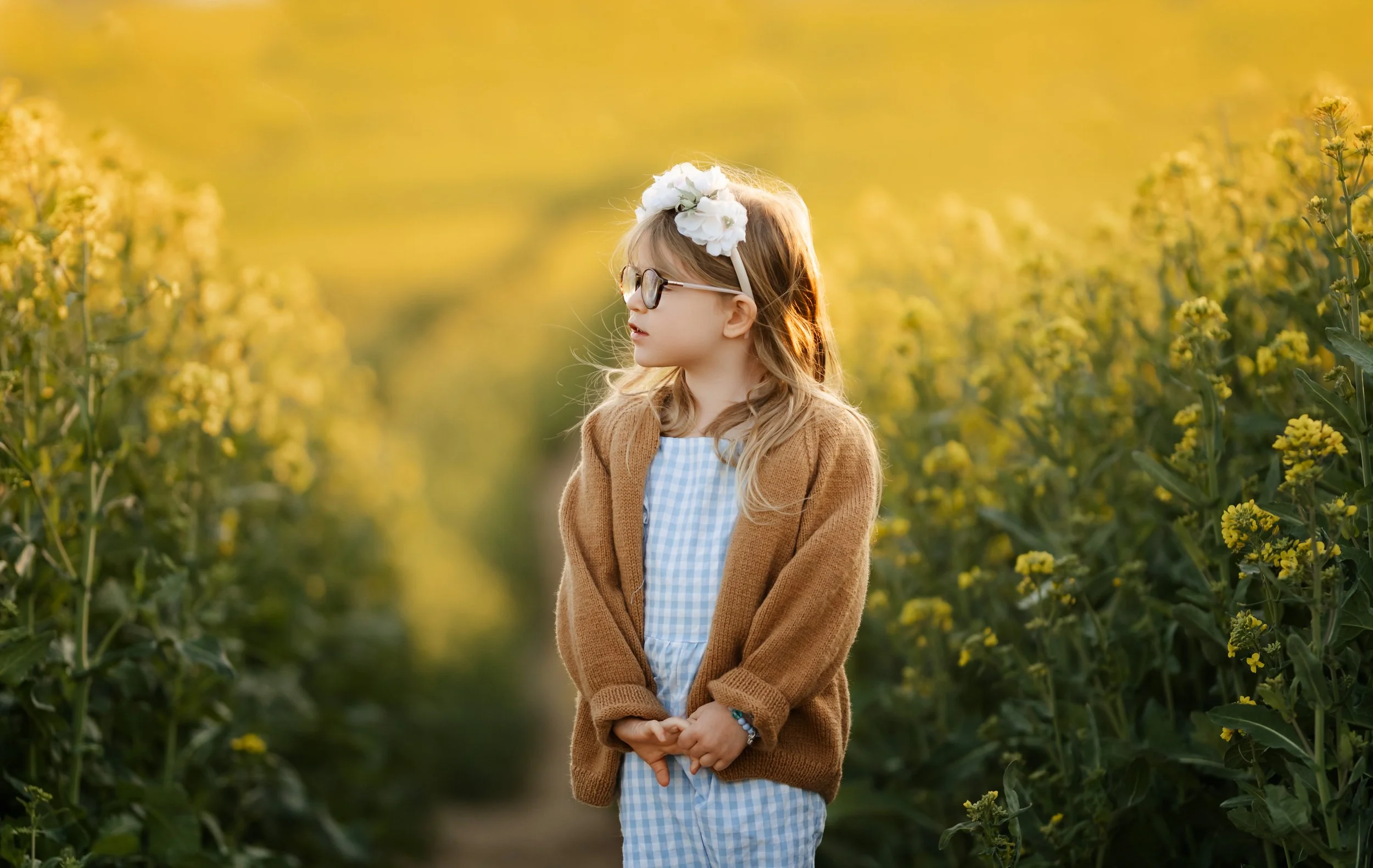 Jeune fille avec lunettes et bandeau de fleurs, portant une robe à carreaux bleus et blancs, et un cardigan marron, debout dans un champ de fleurs jaunes au coucher du soleil.