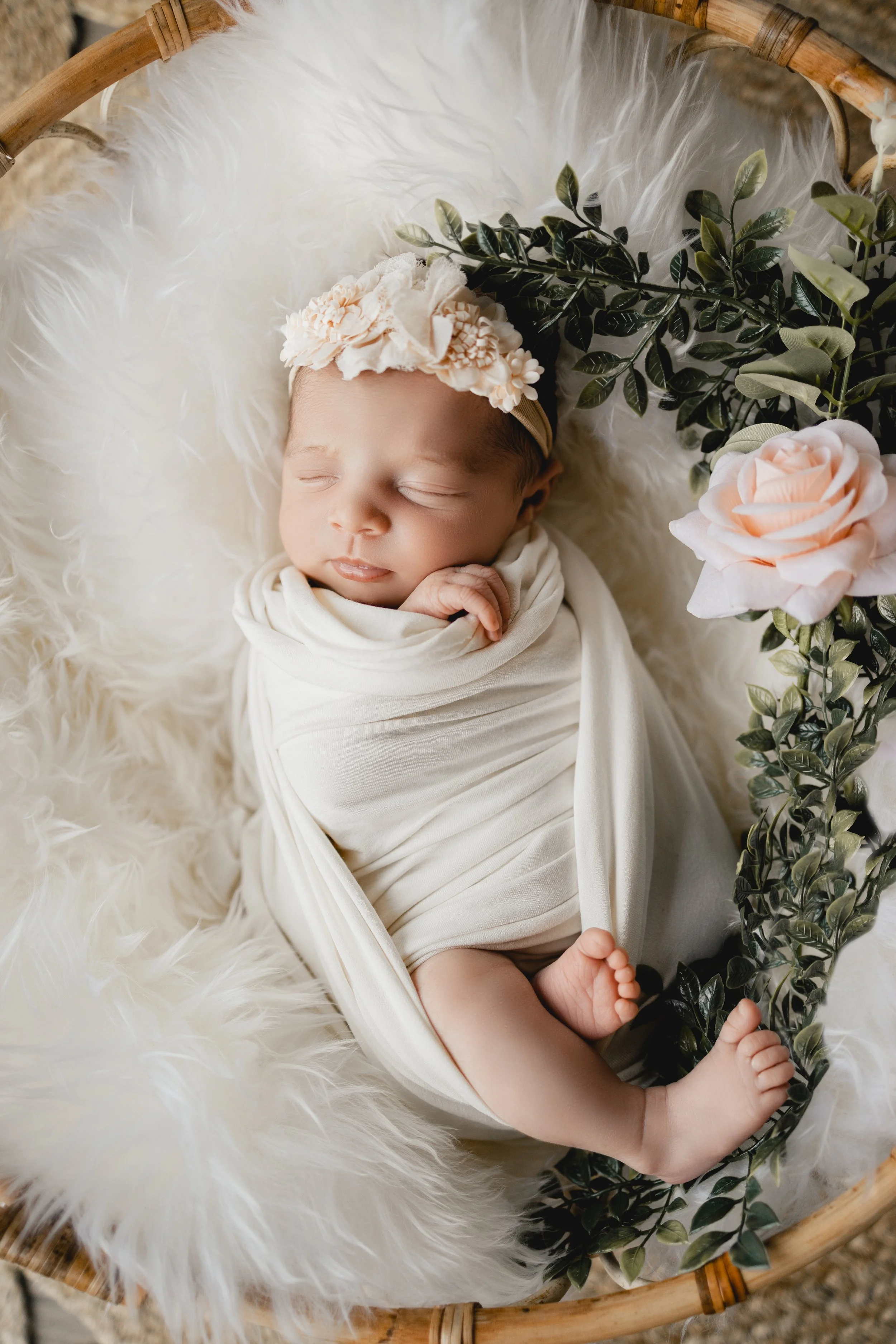Nouveau-né endormi, enveloppé dans un tissu beige, portant un bandeau avec des fleurs, entouré de fleurs et de feuillage, allongé sur une surface en fourrure blanche dans un panier en bois.