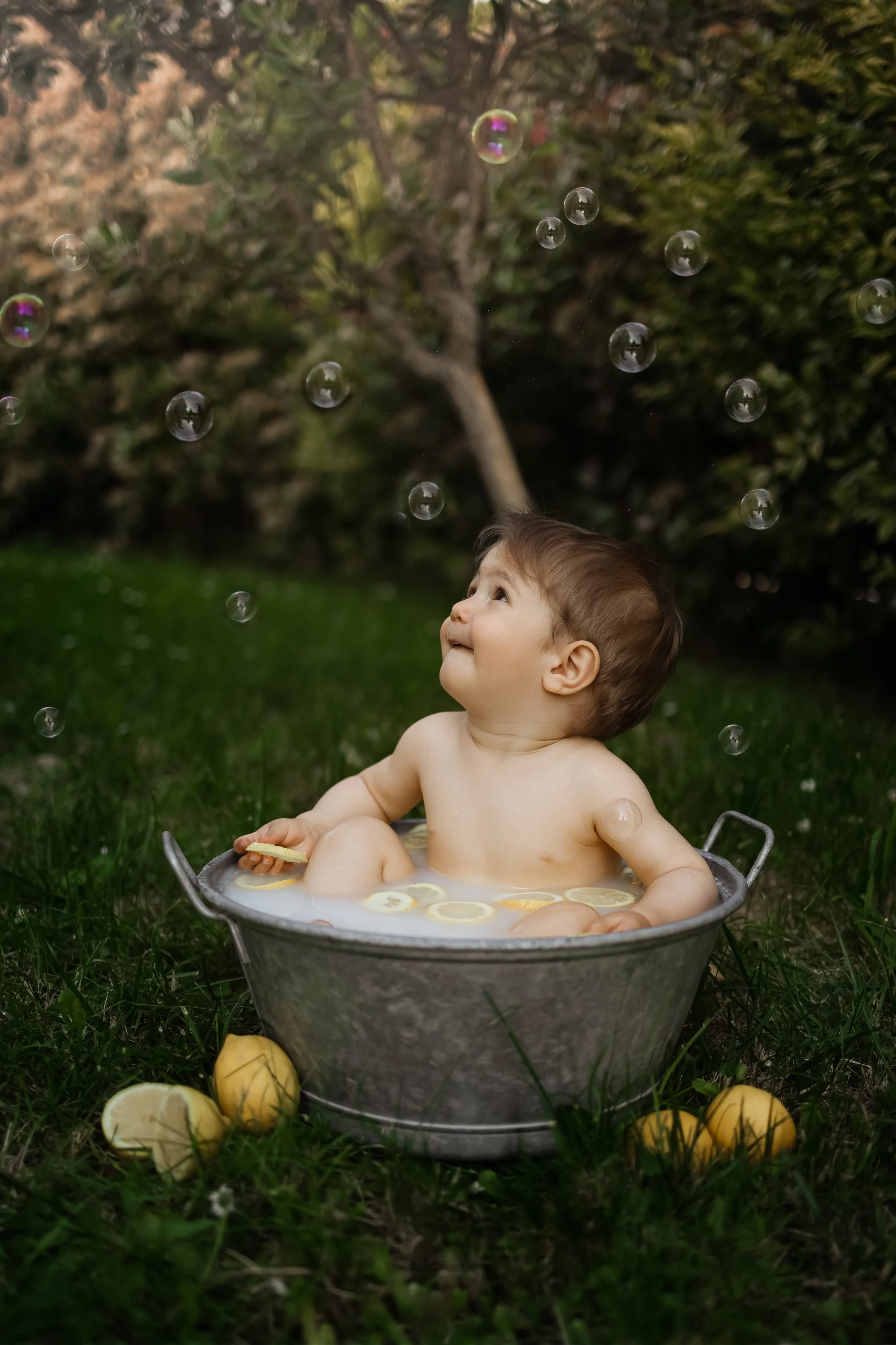 Un bébé assis dans une baignoire en métal rempli de citrons et de citronnade, jouant avec des bulles dans un jardin.
