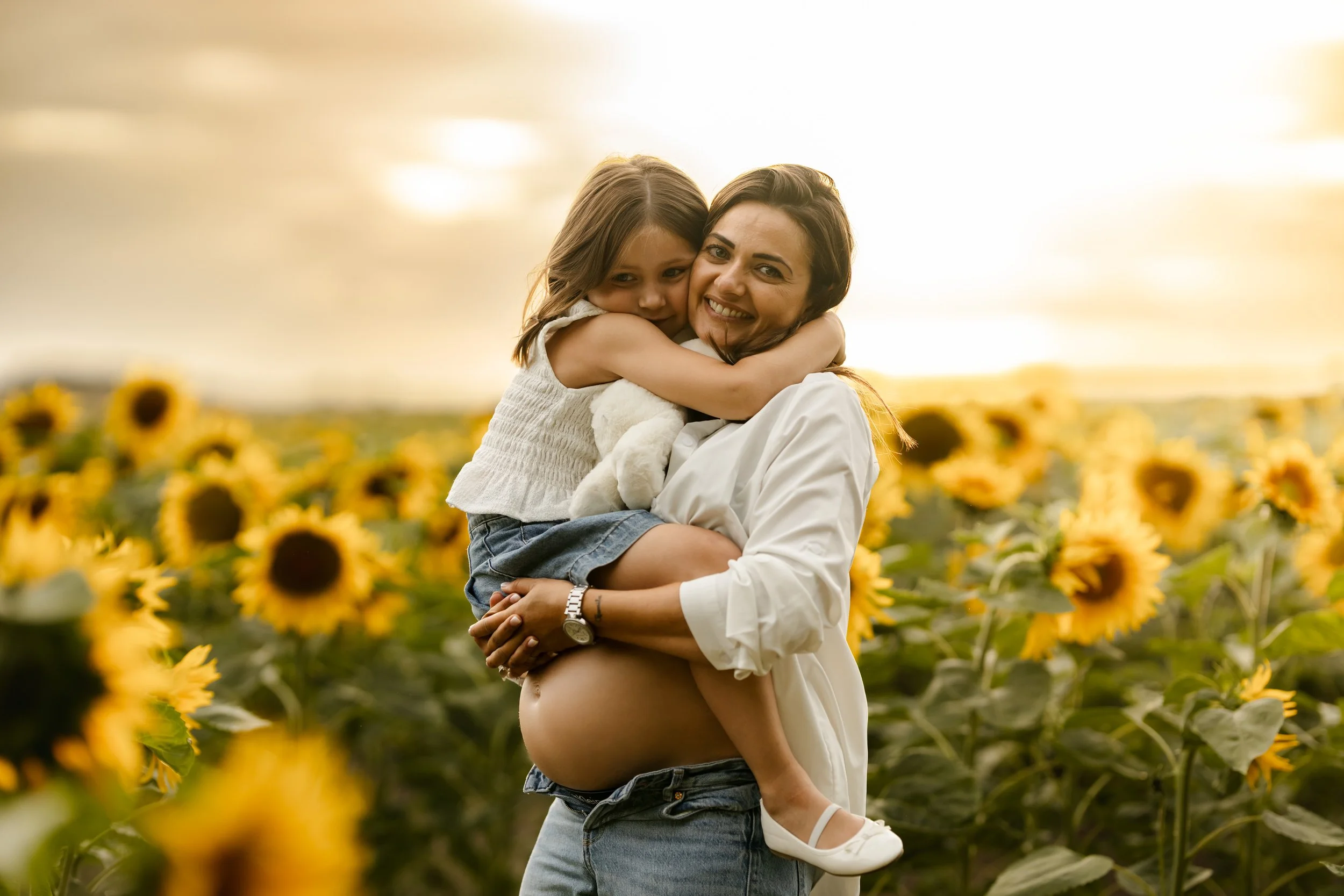 Une femme enceinte avec un petit garçon dans ses bras, tous deux souriants, dans un champ de tournesols au coucher du soleil.