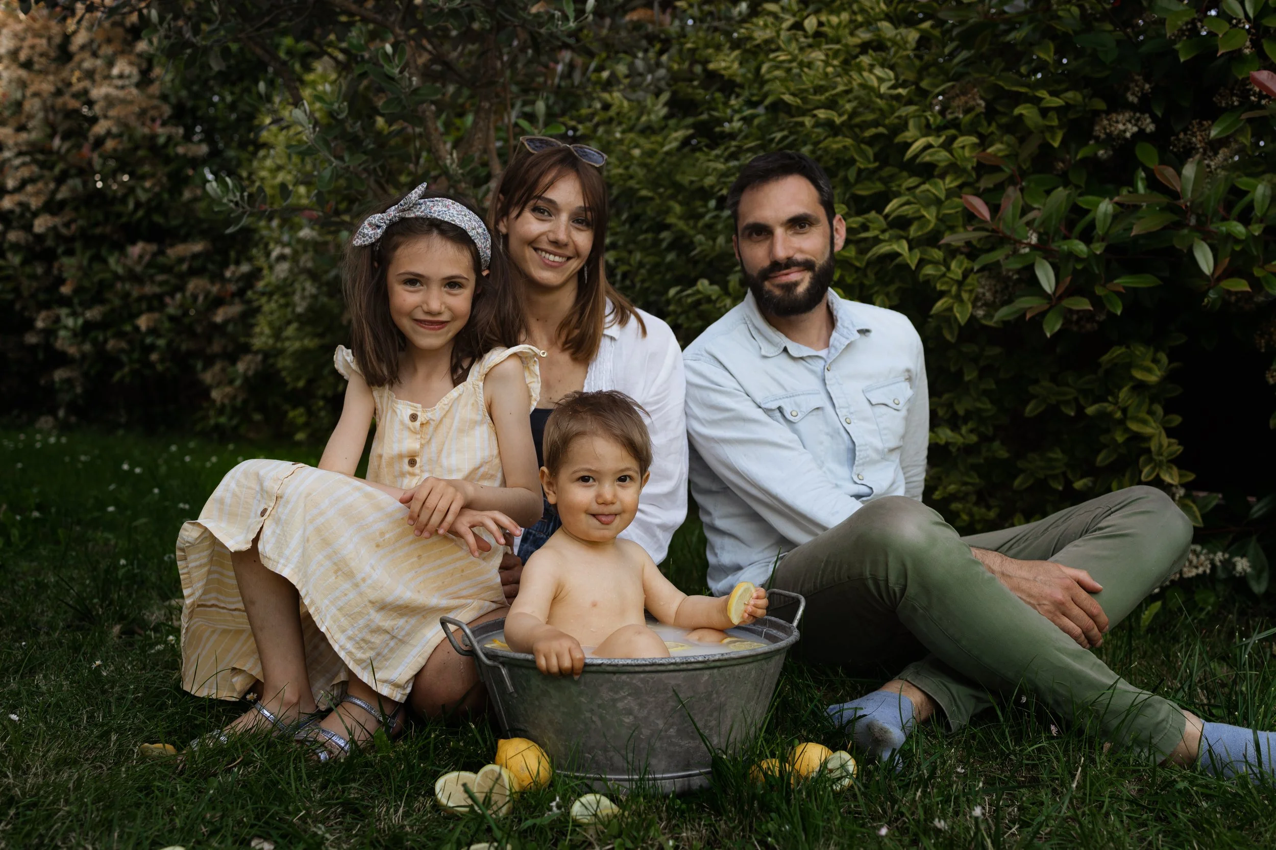 Une famille de cinq personnes, comprenant deux adultes et trois enfants, assis sur l'herbe dans un jardin. Un bébé assis dans un bac rempli d'eau, tenant un citron, les autres enfants sont une fille et un garçon. Les adultes sont derrière eux, souria