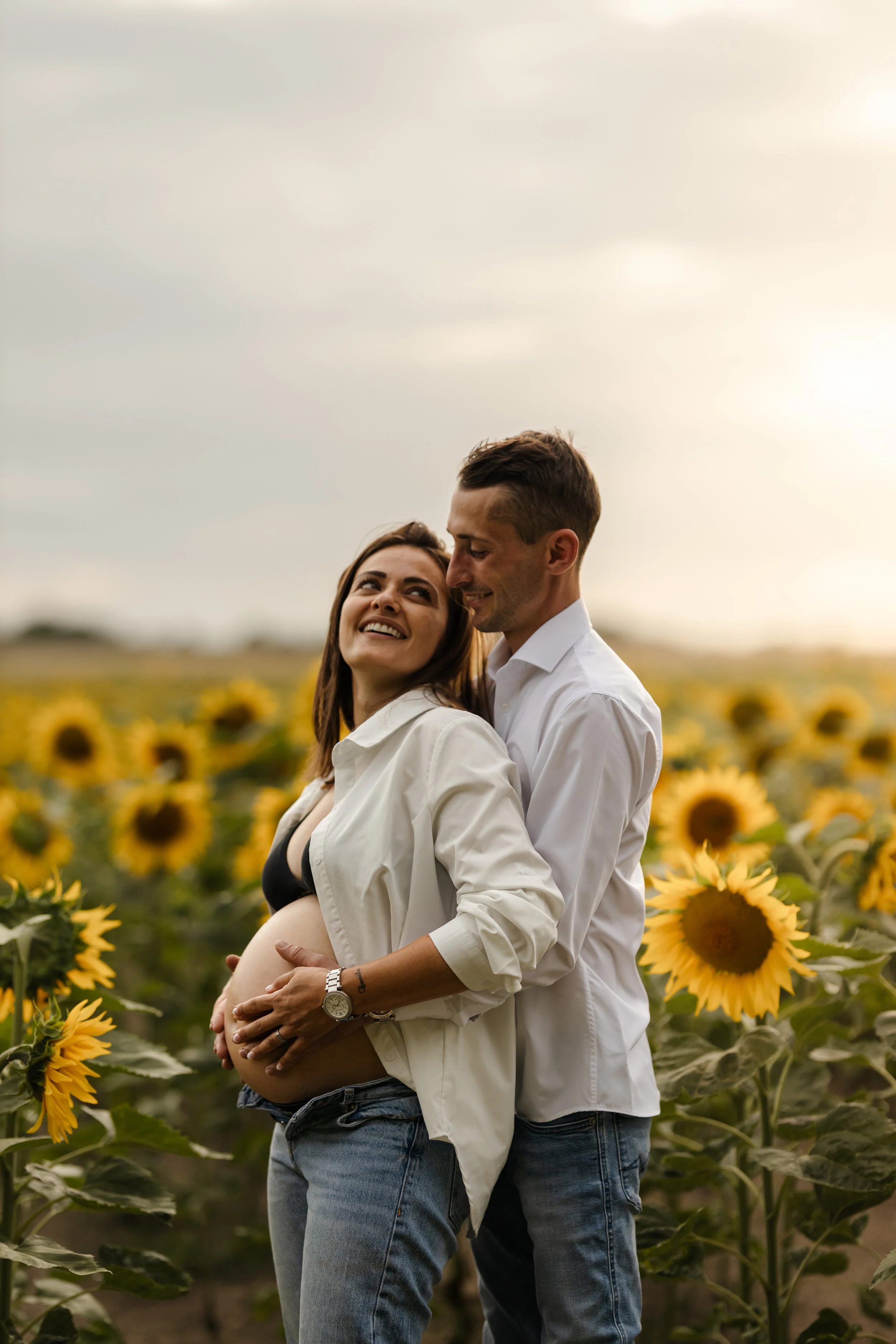 Un couple souriant dans un champ de tournesols, la femme étant enceinte, lors d'une scène de portrait en plein air au coucher du soleil.
