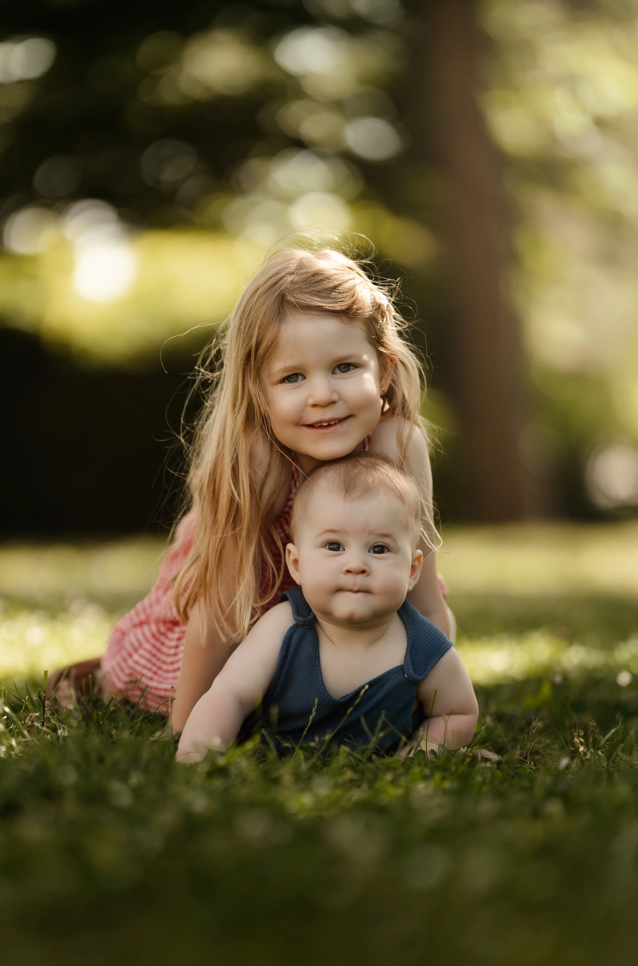 Deux enfants, un garçon plus âgé avec de longs cheveux blonds et une fille plus jeune avec des cheveux courts, jouent sur l'herbe dans un parc ensoleillé.