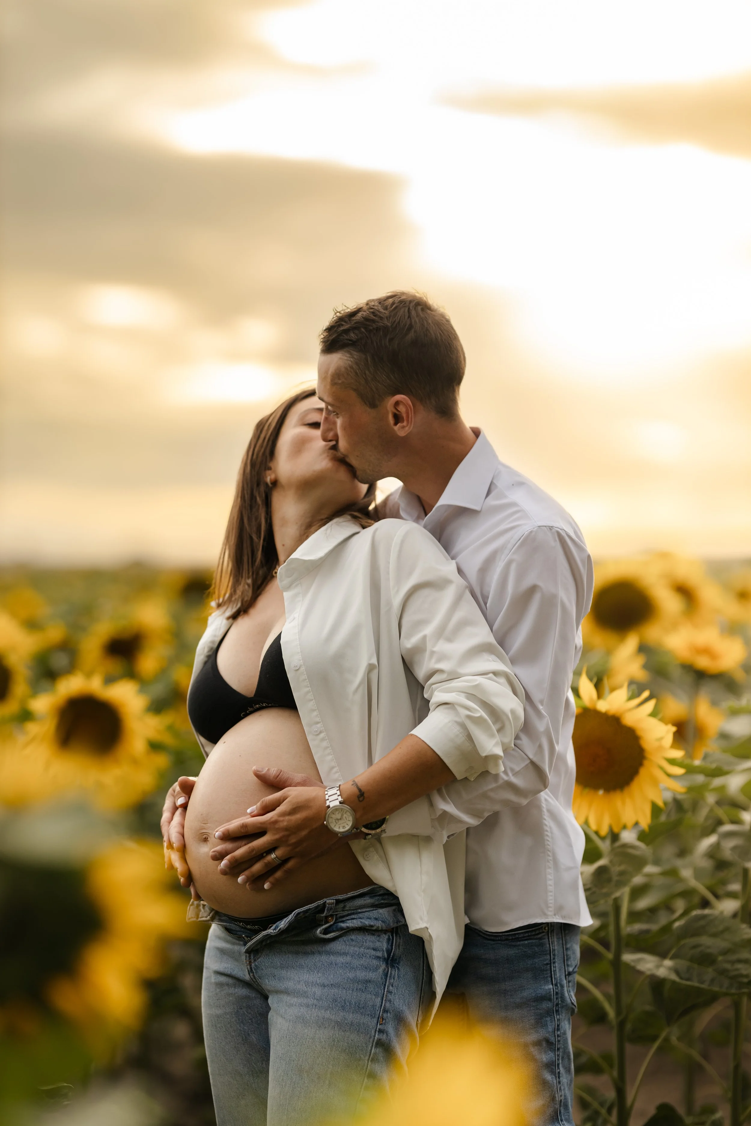 Un couple s'embrasse tendrement dans un champ de fleurs de tournesols, femme enceinte, ciel voilé au coucher du soleil.