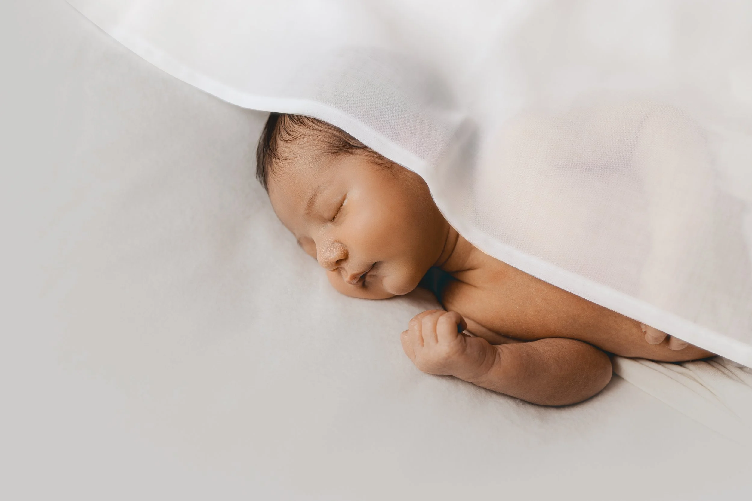 Un bébé endormi sur le côté avec une couverture blanche partiellement couvrante, nu, avec des yeux fermés et des cheveux foncés.