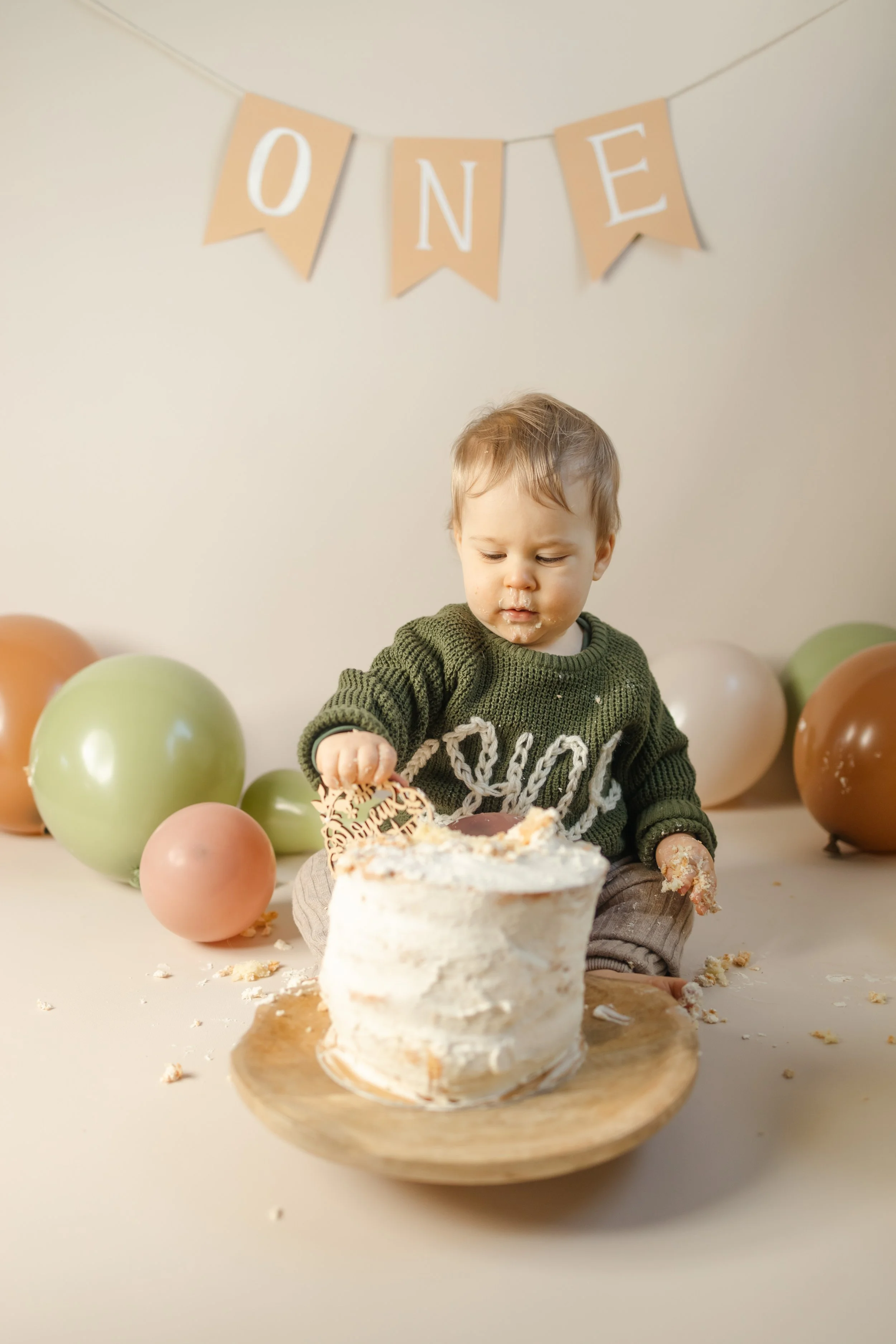 Un bébé assis devant un gâteau d'anniversaire avec des ballons en arrière-plan et une bannière 'ONE' suspendue au plafond.