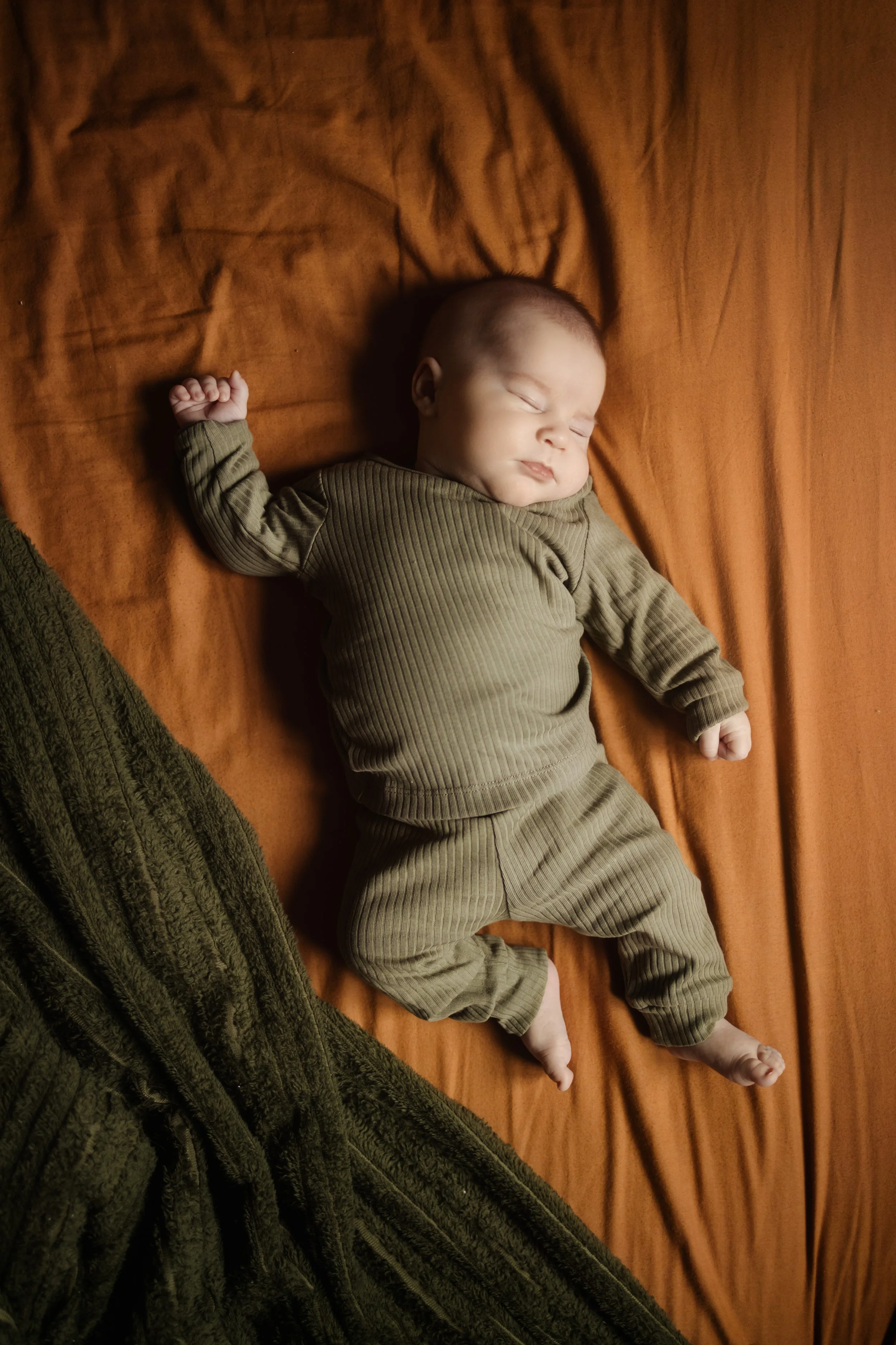 Un bébé qui dort paisiblement sur un lit en tissu marron, habillé d'une tenue marron clair en velours côtelé, avec un motif de rayures. Le bébé est allongé sur le dos, avec une main levée et l'autre repose sur le lit.