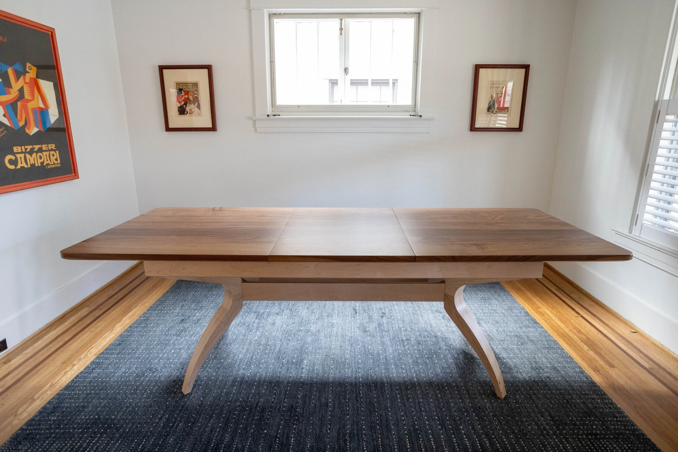 A rectangular wooden table with curved legs in a bright room with white walls, a window, and framed artwork.