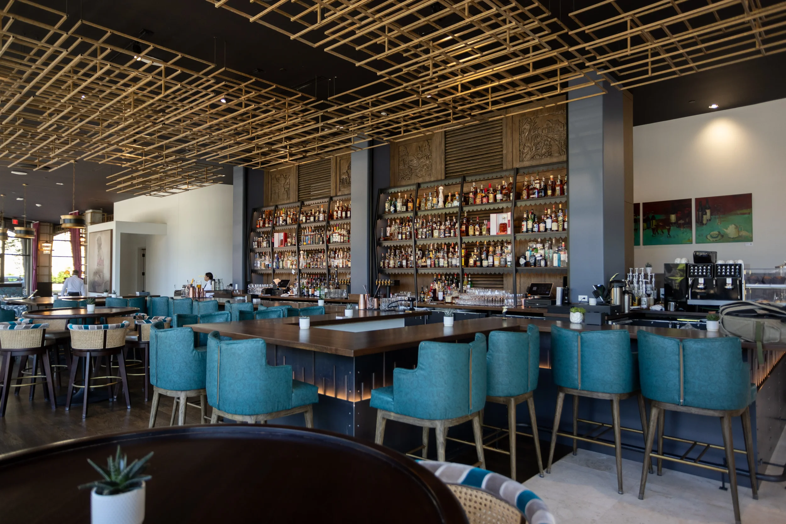 Upscale bar with high ceilings, wood grid ceiling decor, tall shelves of liquor bottles, seafoam green barstools, a few patrons, and a coffee station.