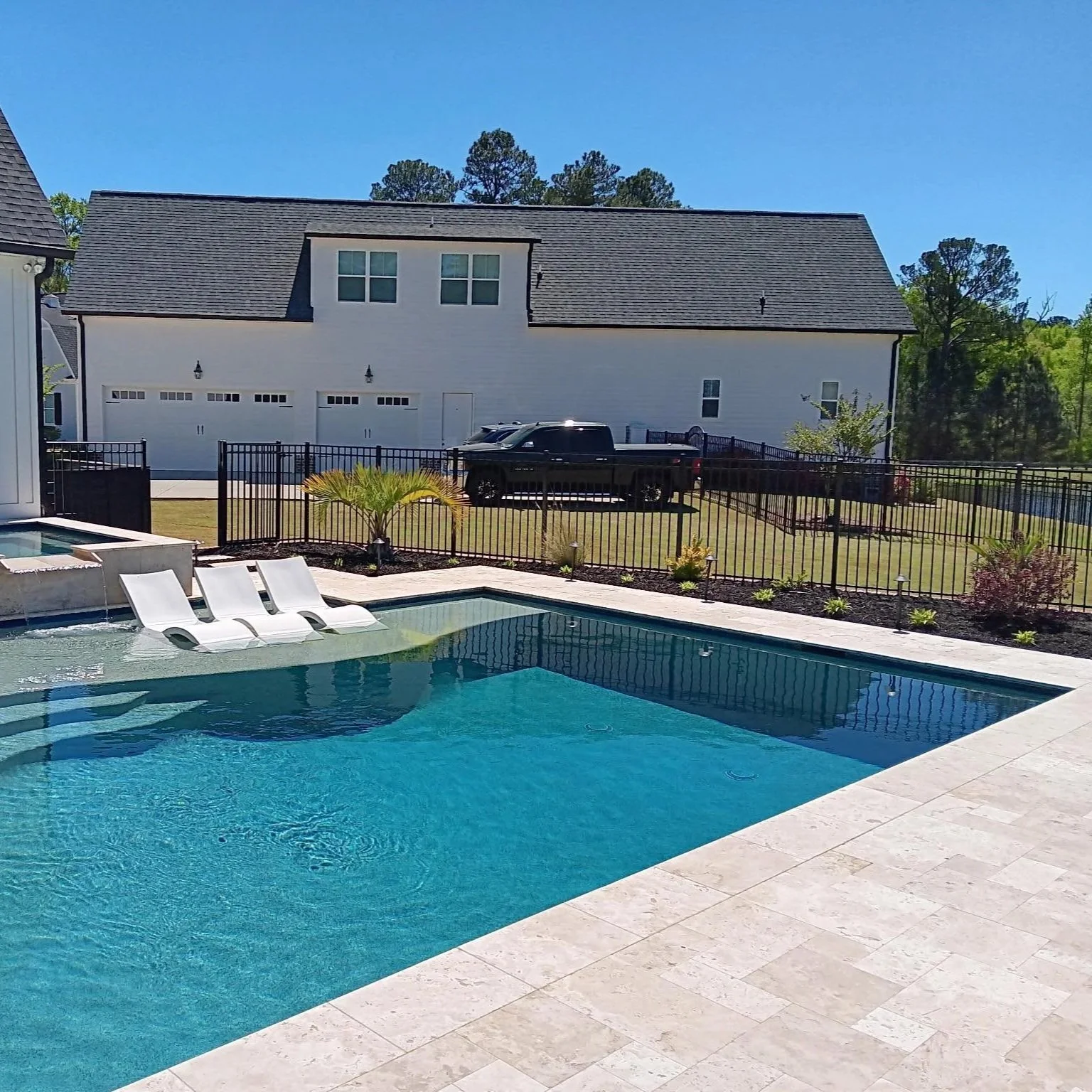 Backyard scene with a swimming pool with clear water, white pool loungers, a small palm tree, a black fence, a house with a large white garage, a black truck, and a bright blue sky.