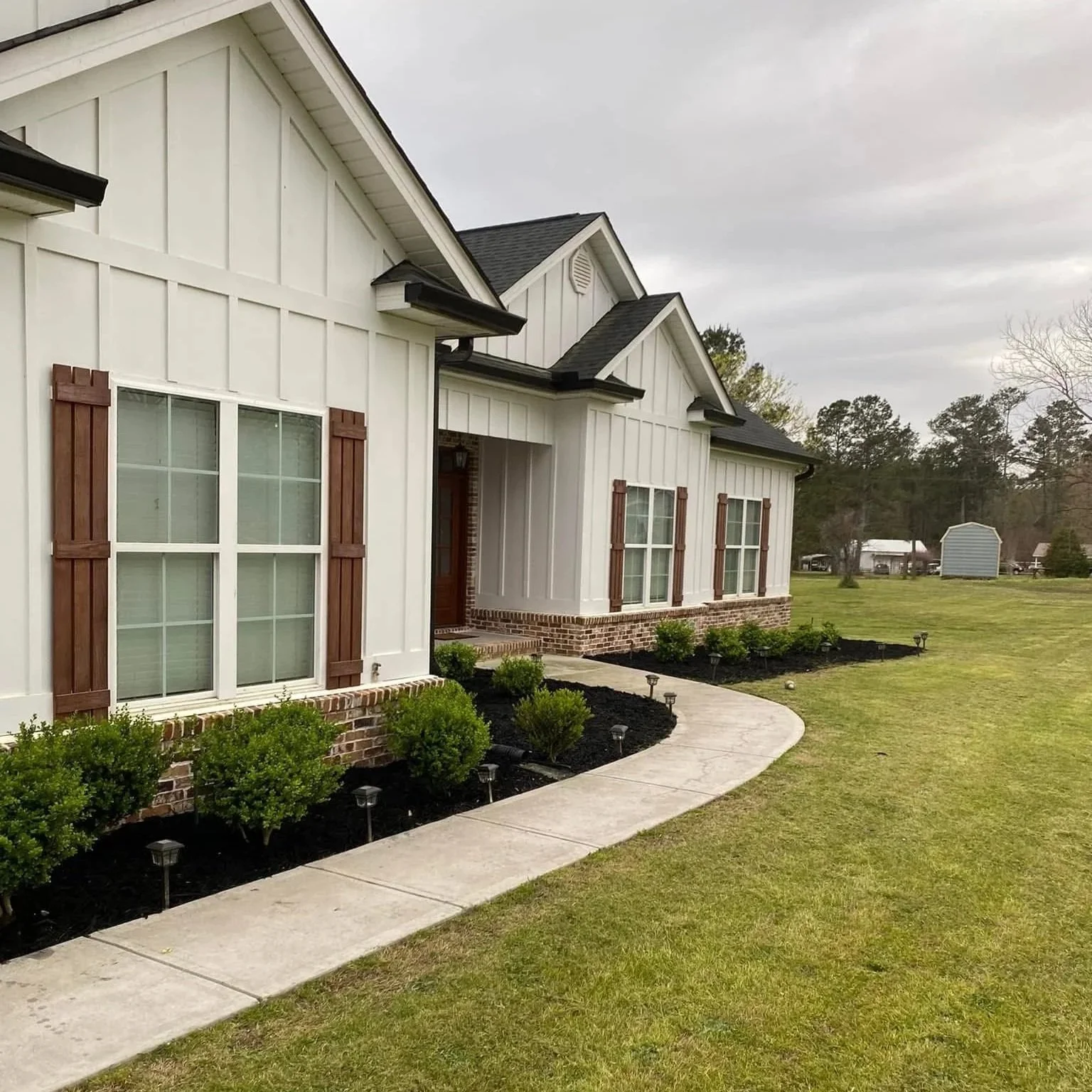 Front view of a white house with brown shutters, a curved concrete walkway, green shrubs, and a well-maintained lawn, with trees and a small shed in the background under an overcast sky.