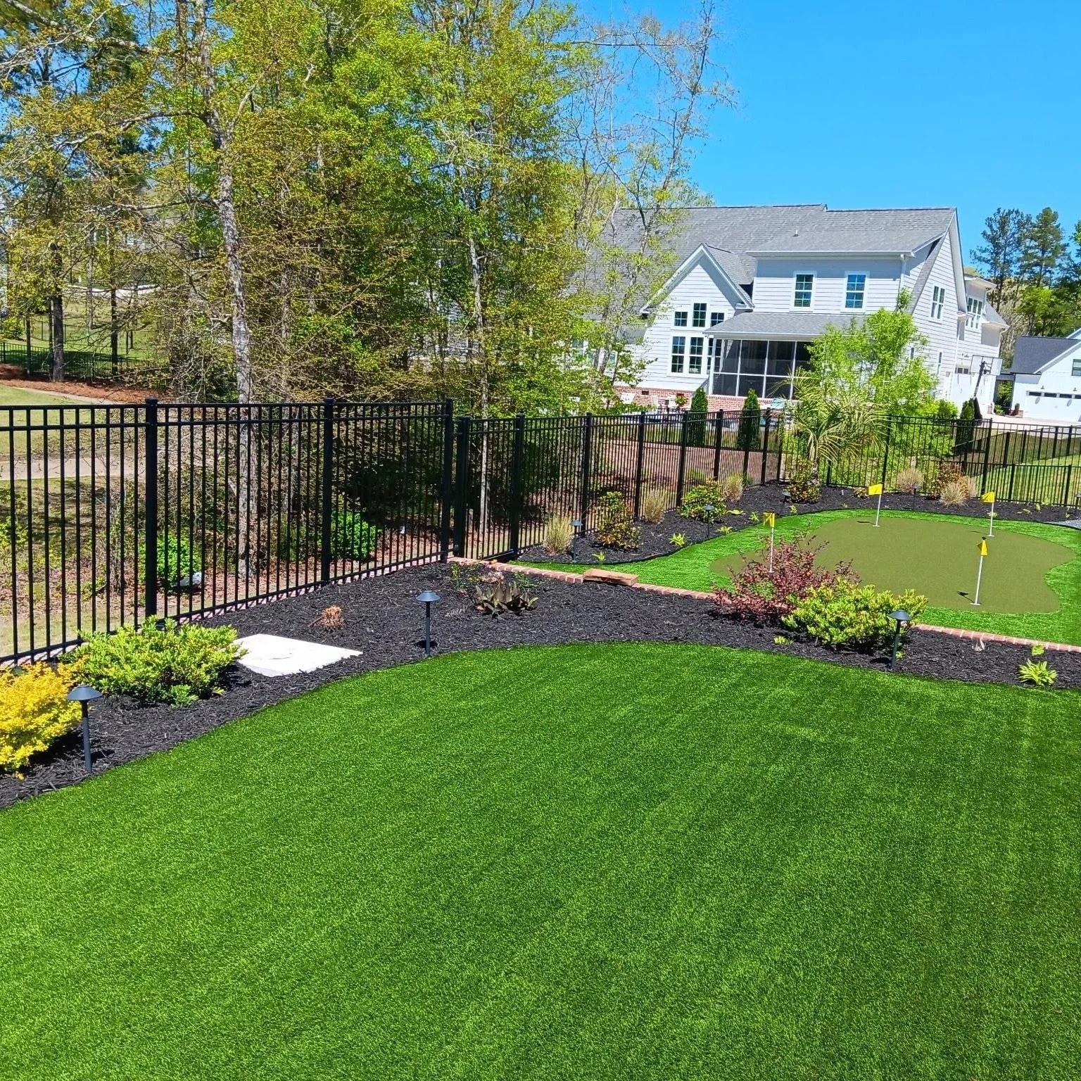 A backyard with a green lawn, a small putting green with four yellow flags, surrounded by landscaping with shrubs, small plants, and garden lights, enclosed by a black metal fence, with a large white house and trees in the background under a clear bl