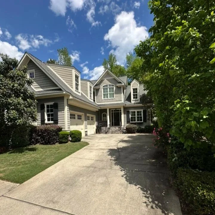 Large gray suburban house with multiple gabled roofs, landscaped front yard, and a concrete driveway leading to a two-car garage, under a partly cloudy blue sky.