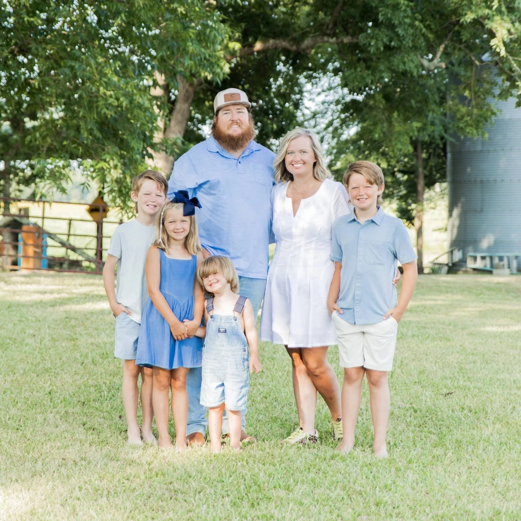 A family of seven posing outdoors, with a man, woman, and five children, on a grassy area with trees and buildings in the background.