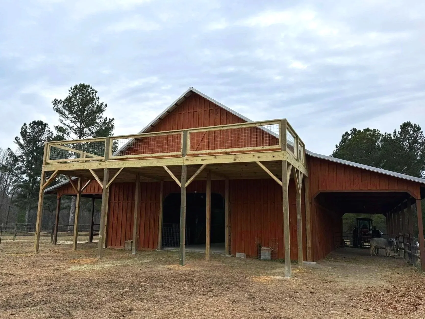 A wooden barn with a second-story balcony, situated on a farm with animals in the background, under a cloudy sky.