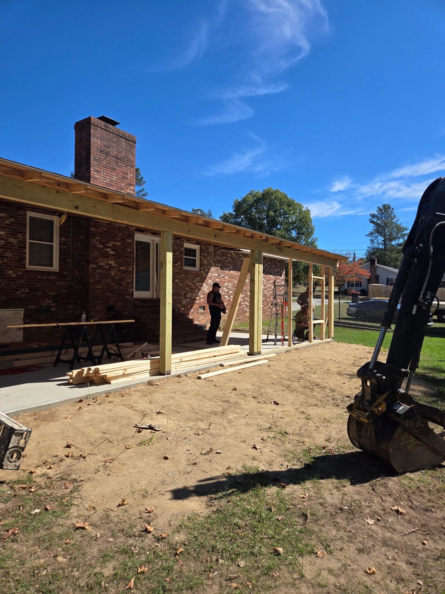 Construction of a wooden porch or patio extension at a brick house under sunny skies. Construction workers are on site, with tools and materials in use.