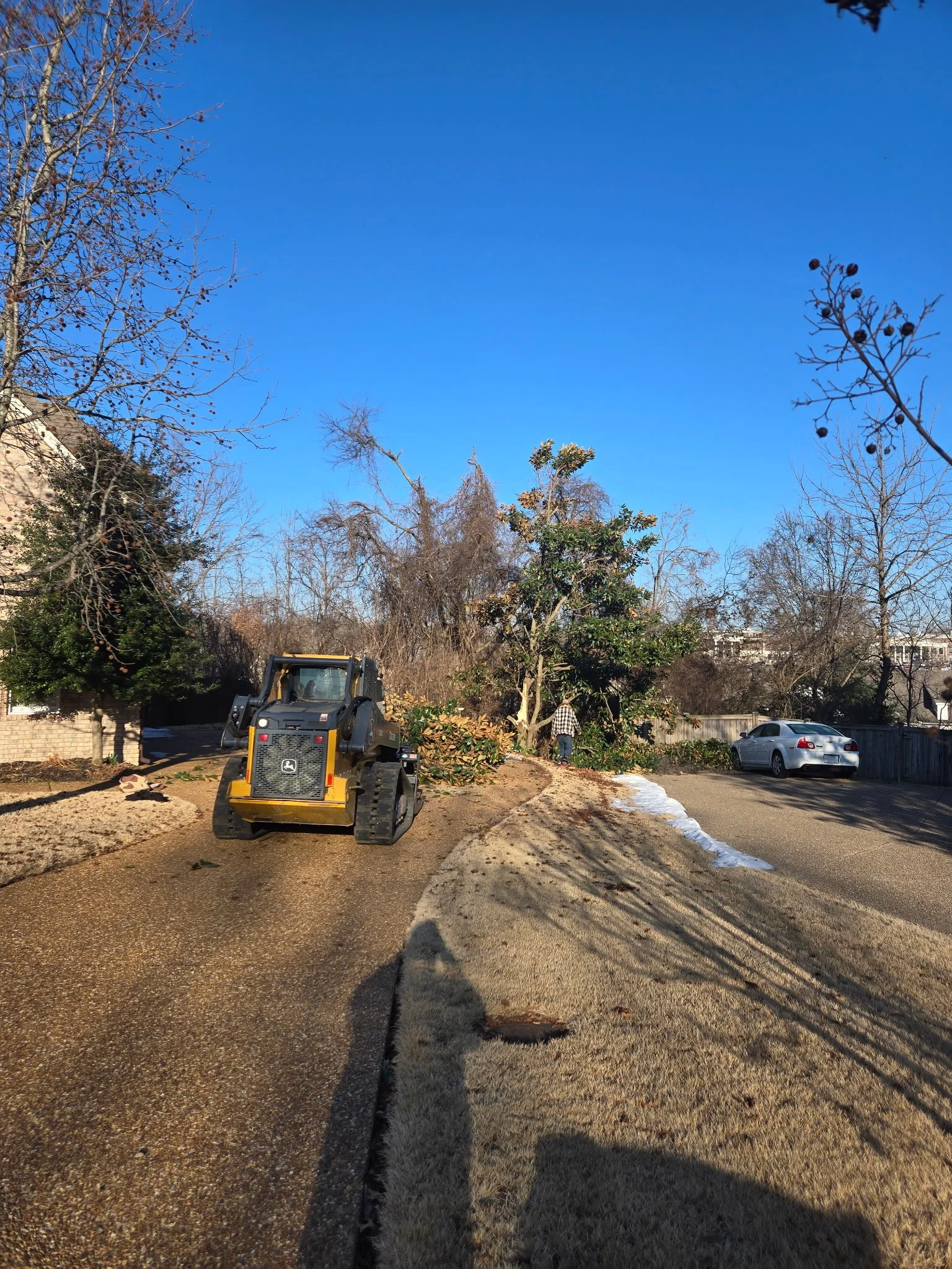 A backyard scene with a small bulldozer parked on a gravel path, leafless trees, a person near a shrub, a silver car, and a clear blue sky.