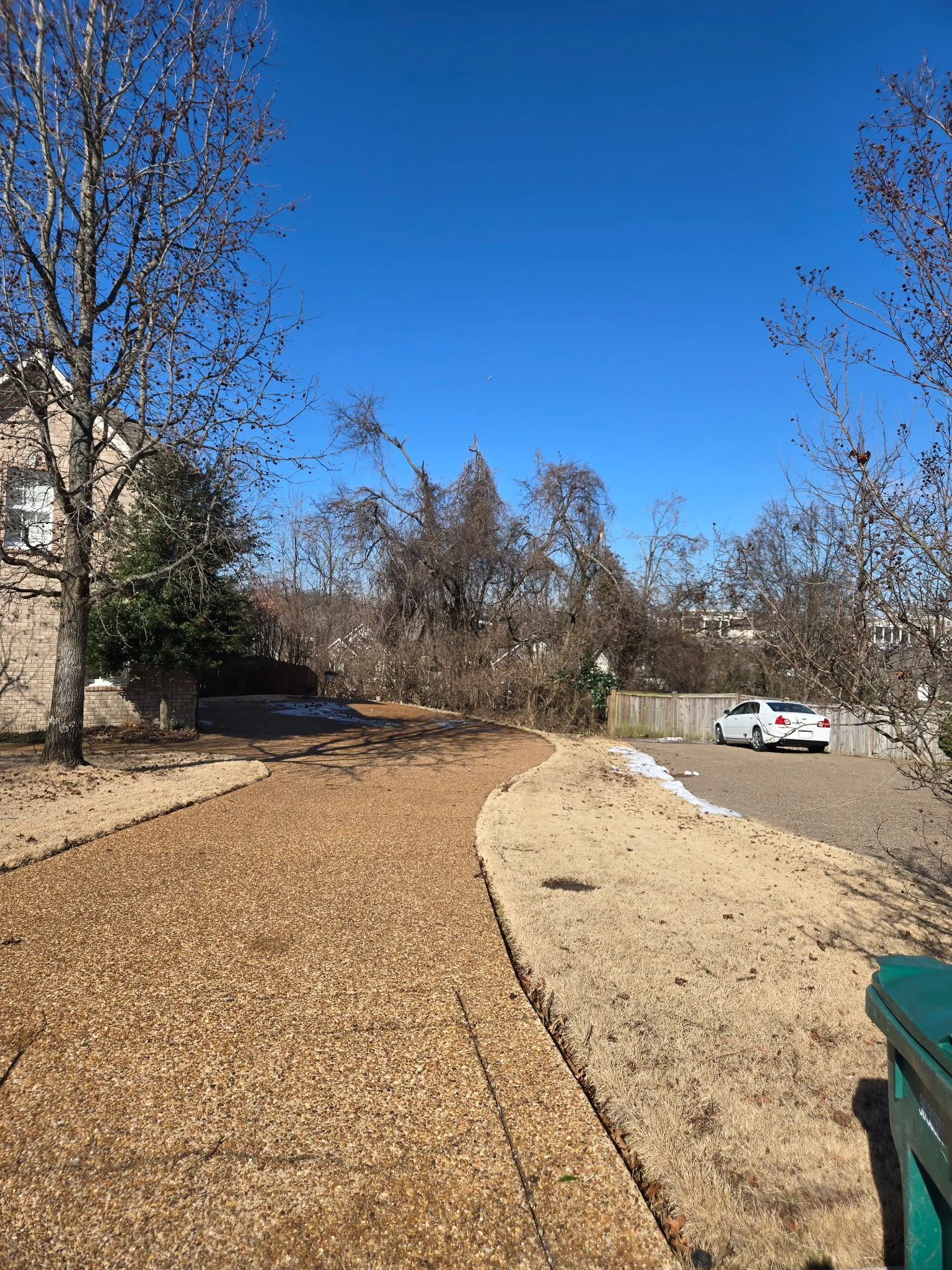 A clear blue sky over a suburban street with a sidewalk, some snow patches, leafless trees, a white car parked on a driveway, and a green trash bin on the right.