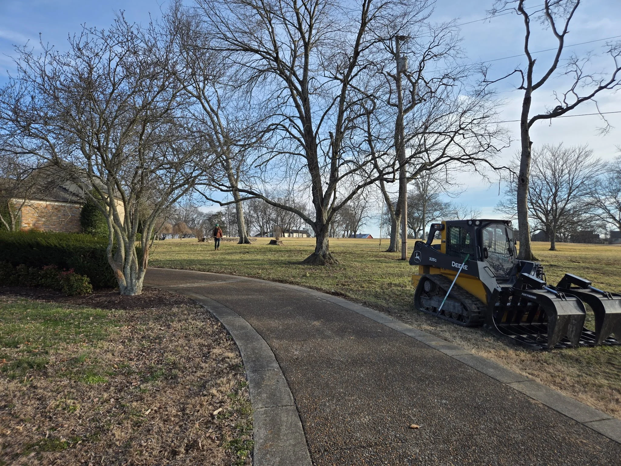 A park with leafless trees, a gravel path, and a John Deere skid-steer loader parked on the grass. A person is walking in the distance under a partly cloudy sky.