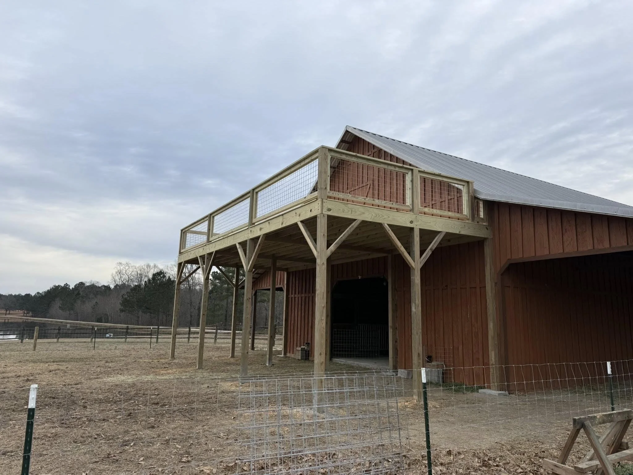 A wooden barn with an upper balcony and railing, situated in a rural area with a fenced pasture and a pond in the background.