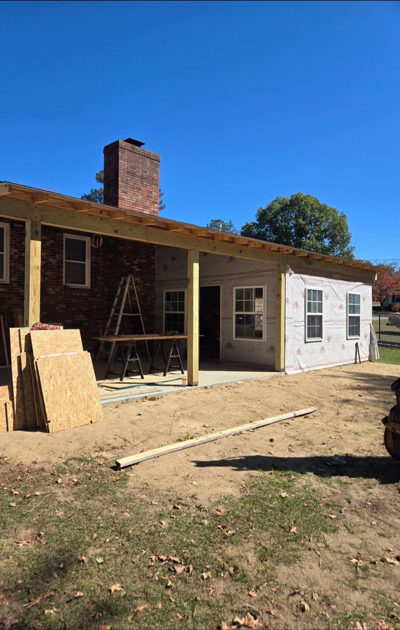 Construction site of a house extension with wooden framework and house wrap siding, tools and materials on site, autumn leaves on the ground, a bright blue sky.