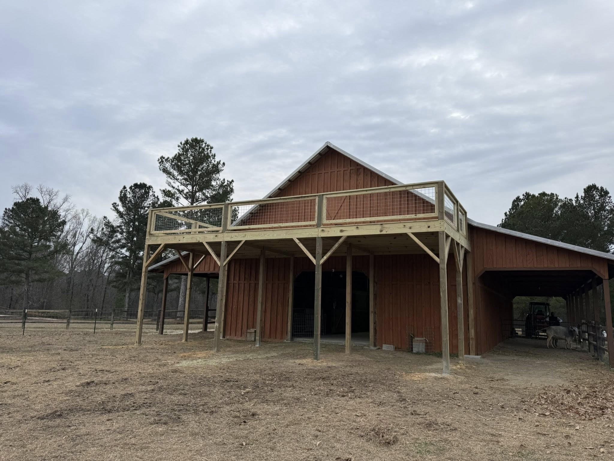 Wooden barn with a second-level outdoor deck, surrounded by trees on a cloudy day.
