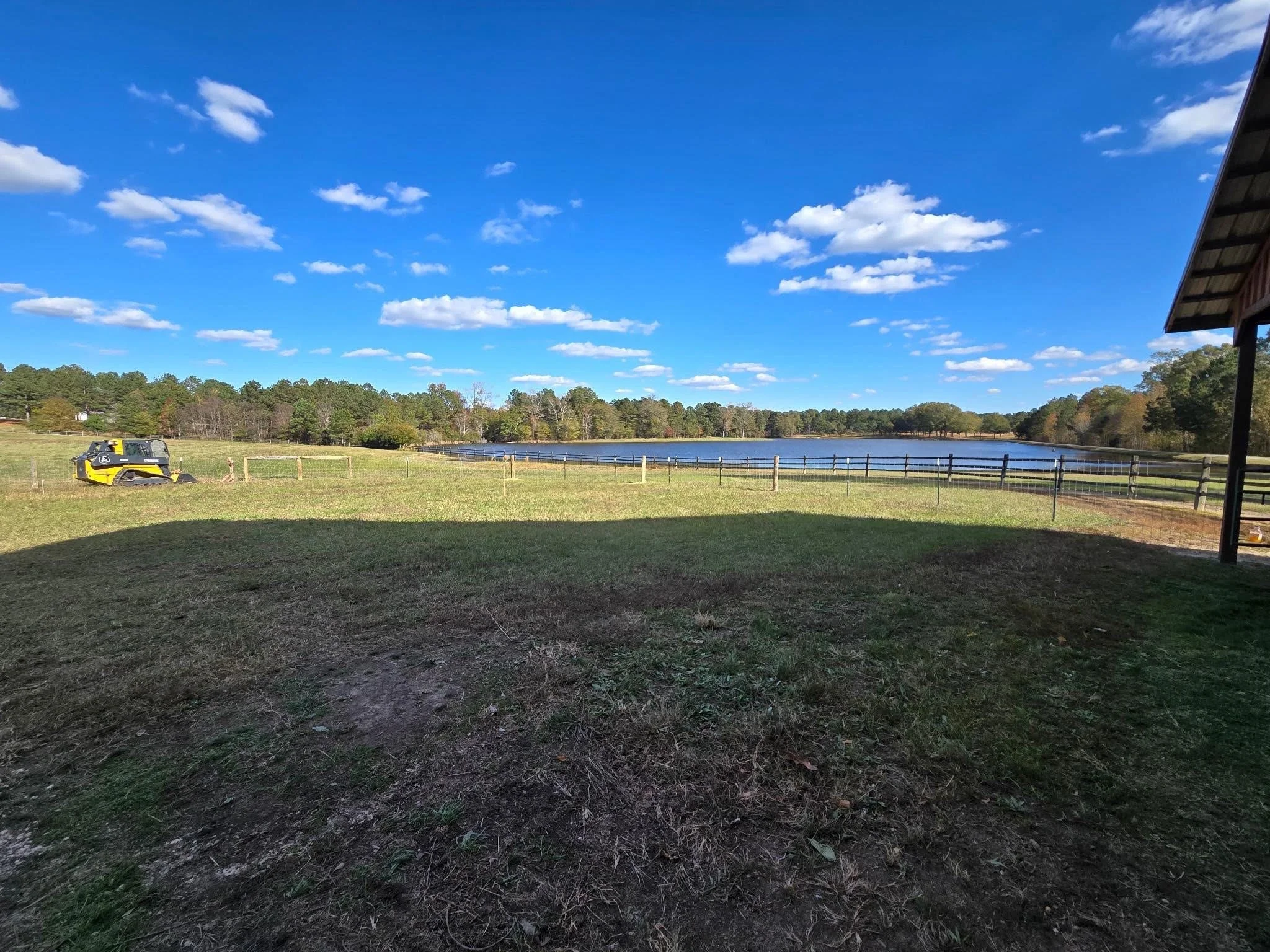 A scenic outdoor view of a grassy field with a pond or lake in the distance, surrounded by trees. A small construction vehicle is parked on the left side. The sky is clear and blue with scattered white clouds, and part of a building's roof is visible
