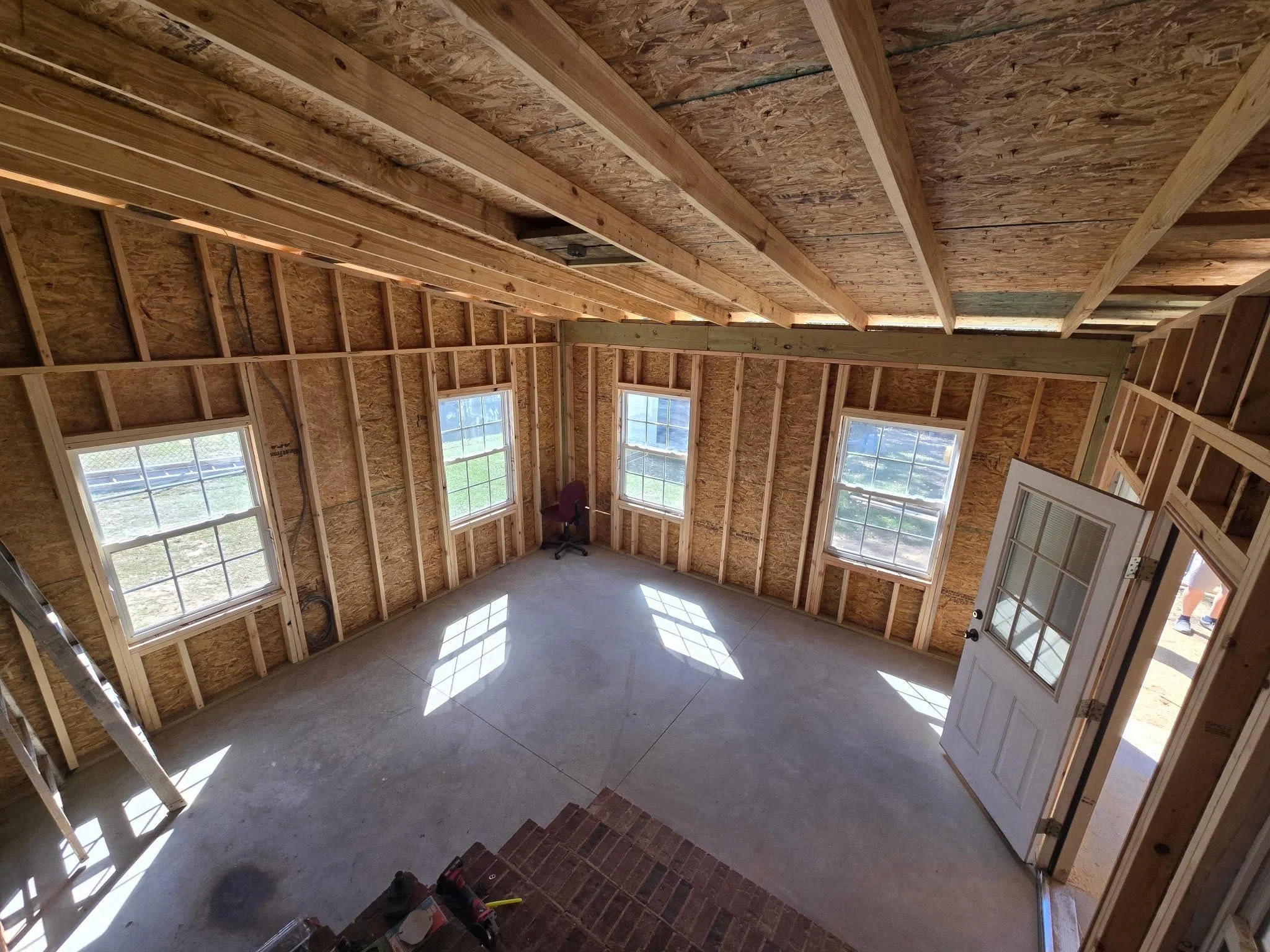 Interior of a house under construction with exposed wooden framing, four windows, an open door, a ladder, and construction tools.