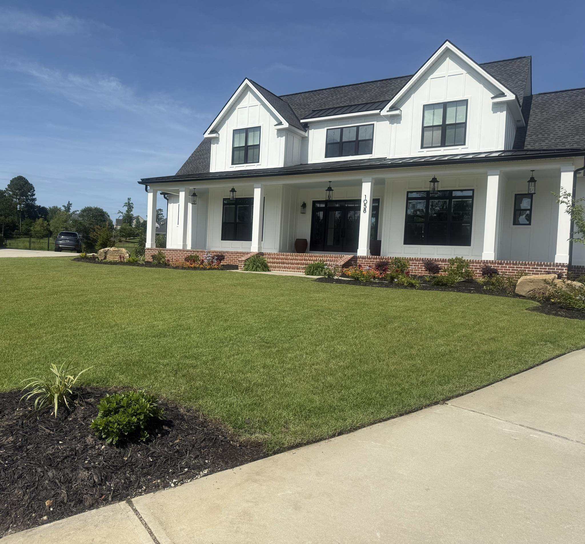 A large white house with black window frames, a brick front porch, and a dark shingle roof, surrounded by a well-manicured lawn and garden beds under a clear blue sky.