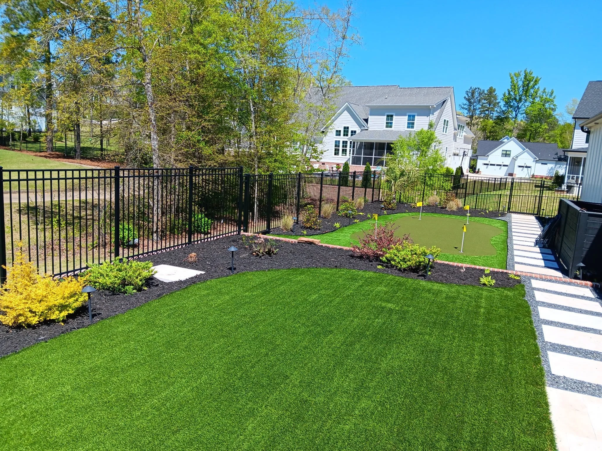 Backyard with well-manicured green grass, a small artificial putting green with holes and flags, black metal fence, stone pathway along fence, landscaped garden beds with small plants and outdoor lights, and neighboring houses under a clear blue sky.