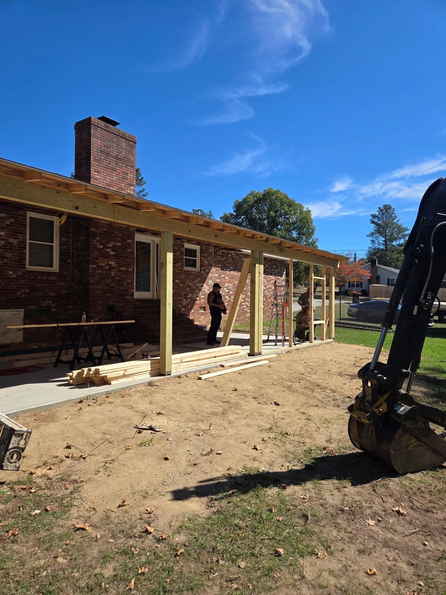 Construction workers building a wooden porch addition on a brick house under a clear blue sky, with construction tools and materials visible.