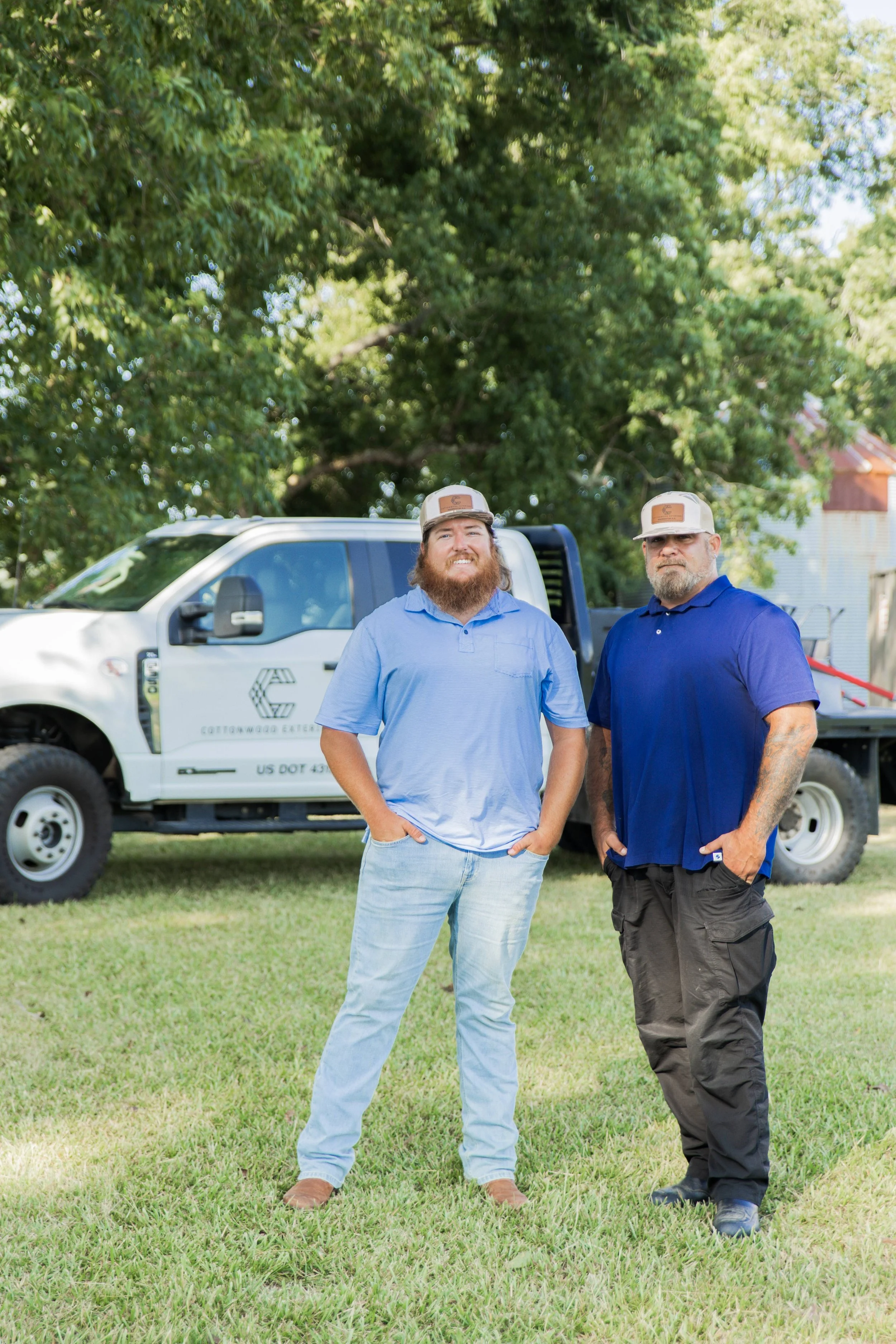 Two men standing outdoors in front of a white utility truck with a large tree in the background.