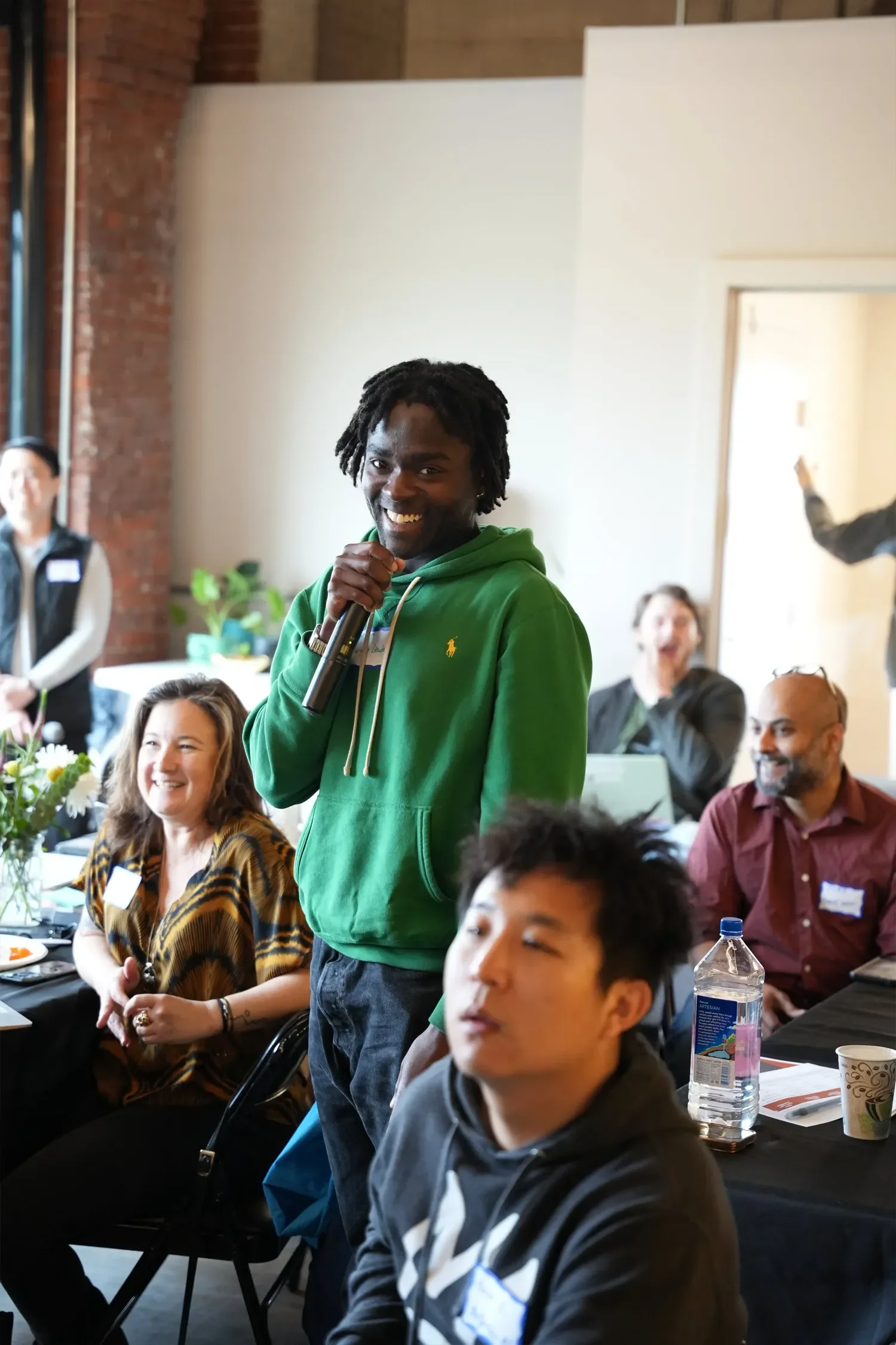 A young man with dark skin and dreadlocks, wearing a green hoodie and holding a microphone, smiling and speaking at a group event. The group is seated at a table in a well-lit room with brick walls, and other attendees are visible, looking engaged.
