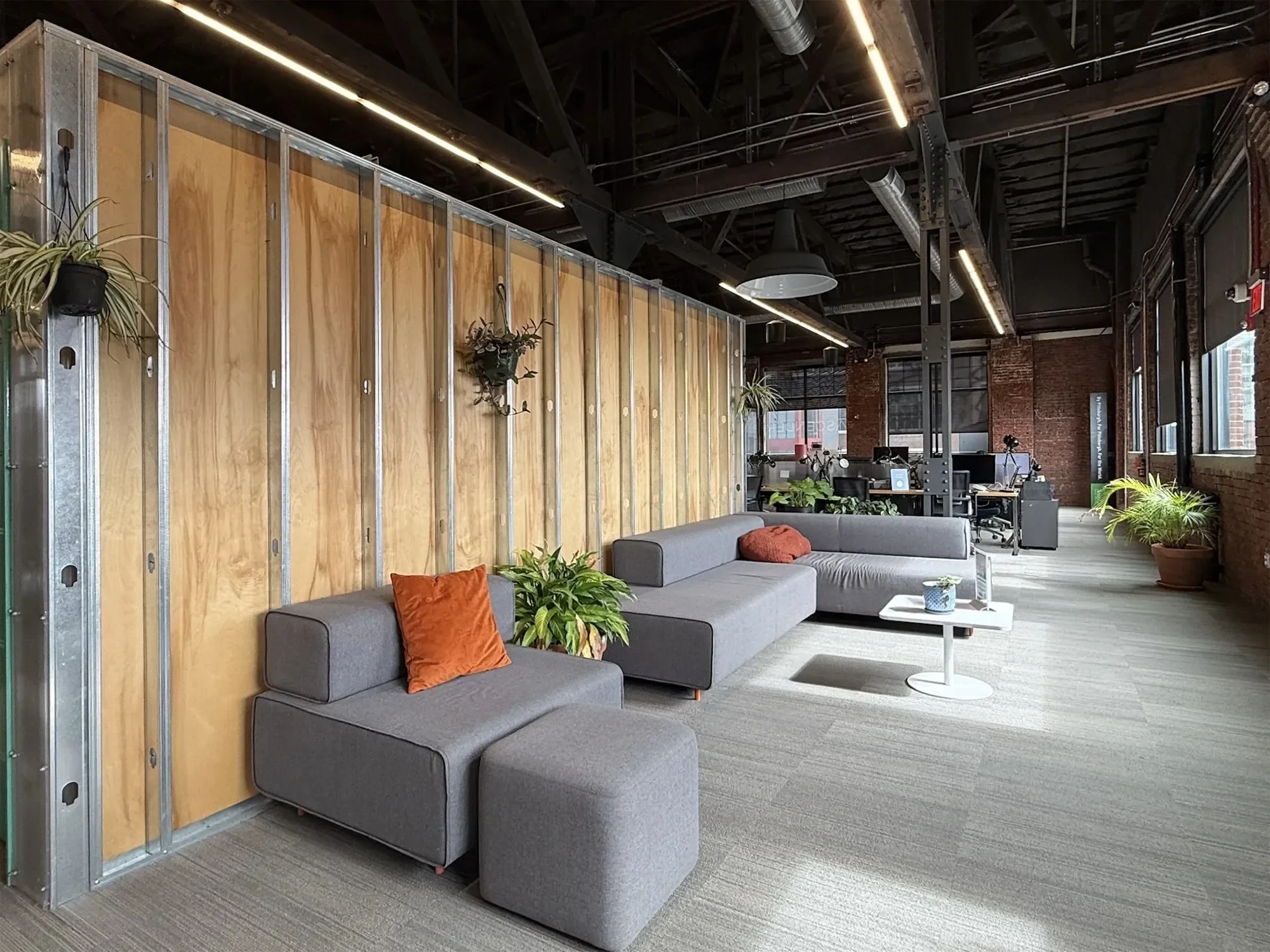 A photo of Ascender's rear flex lounge with gray sectional sofa, orange pillows, potted plants, wooden and metal partition, and workspaces with computers and desks in the background.