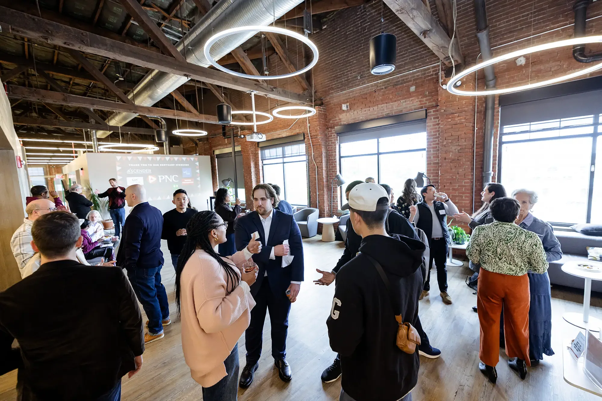 A networking event or conference scene inside a brick-walled industrial-style space with large windows, where diverse people are engaged in conversations and networking, with a projection on the wall displaying logos and a thank you message.