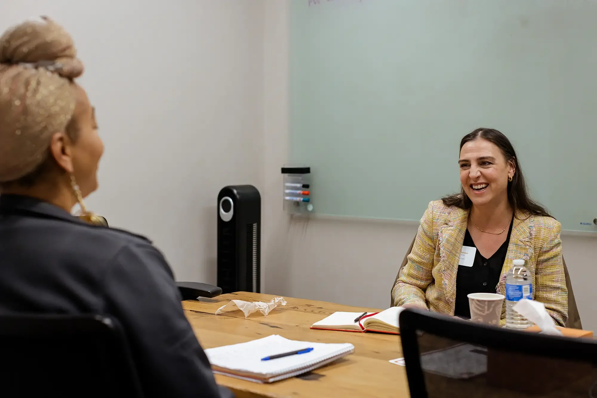 Two women having a conversation in a meeting room, both smiling. One woman, with brown hair, is wearing a plaid blazer and gold earrings, and the other woman, with blonde and glittery hair, is facing away from the camera. The table has notebooks, pen