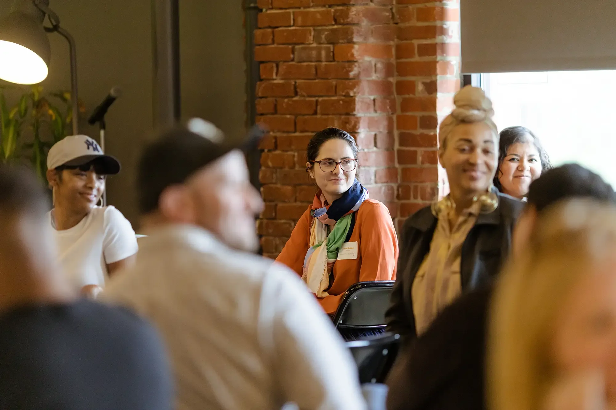A group of diverse people seated in a room with brick walls, attending a gathering or meeting. One woman in the center wears glasses and a colorful scarf, while others are smiling and listening attentively.